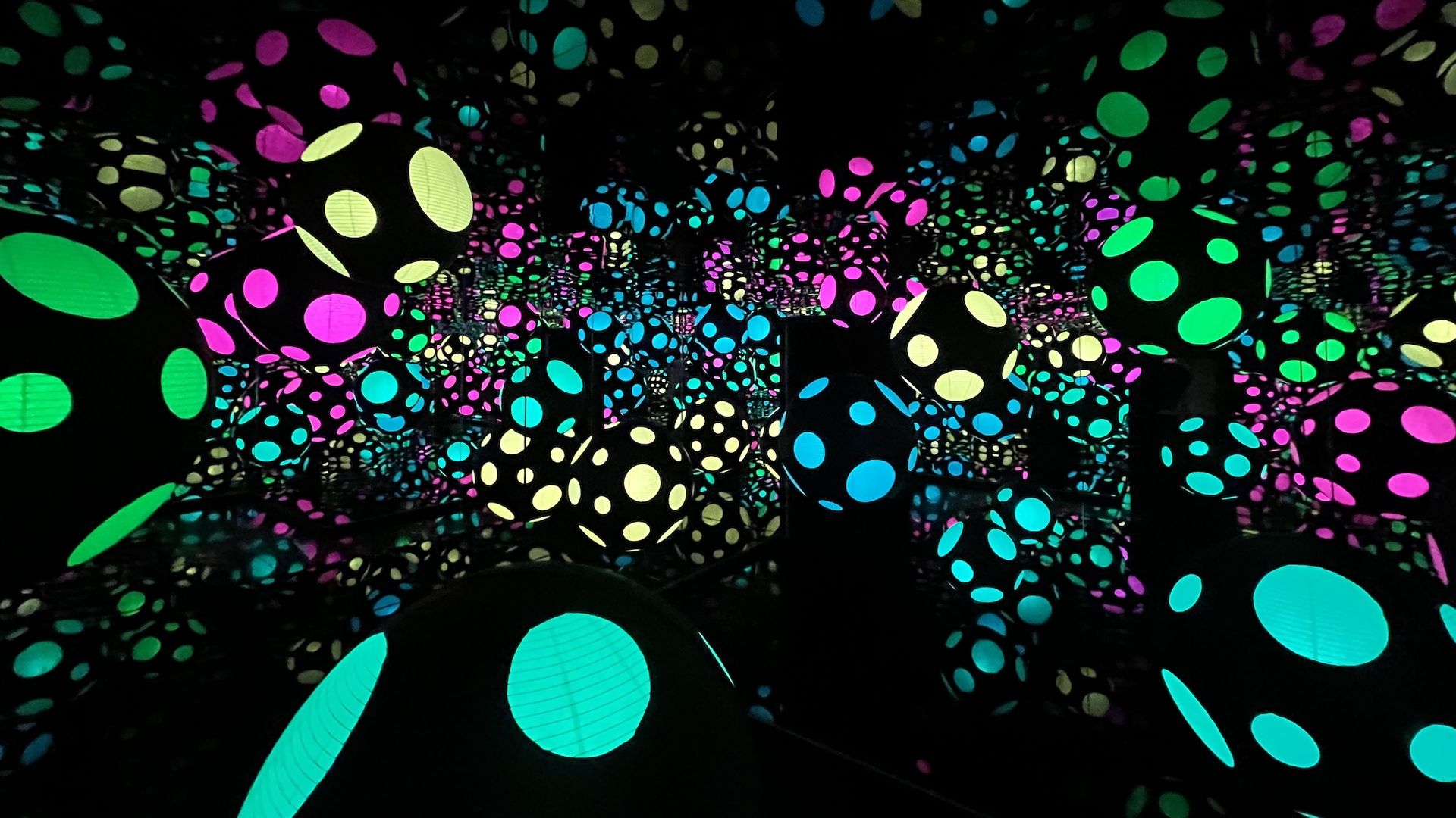 Dark room filled with illuminated multicolored neon dots and oval shapes on mirrored surfaces, creating a tunnel of glowing spheres in green, pink, blue, and yellow.