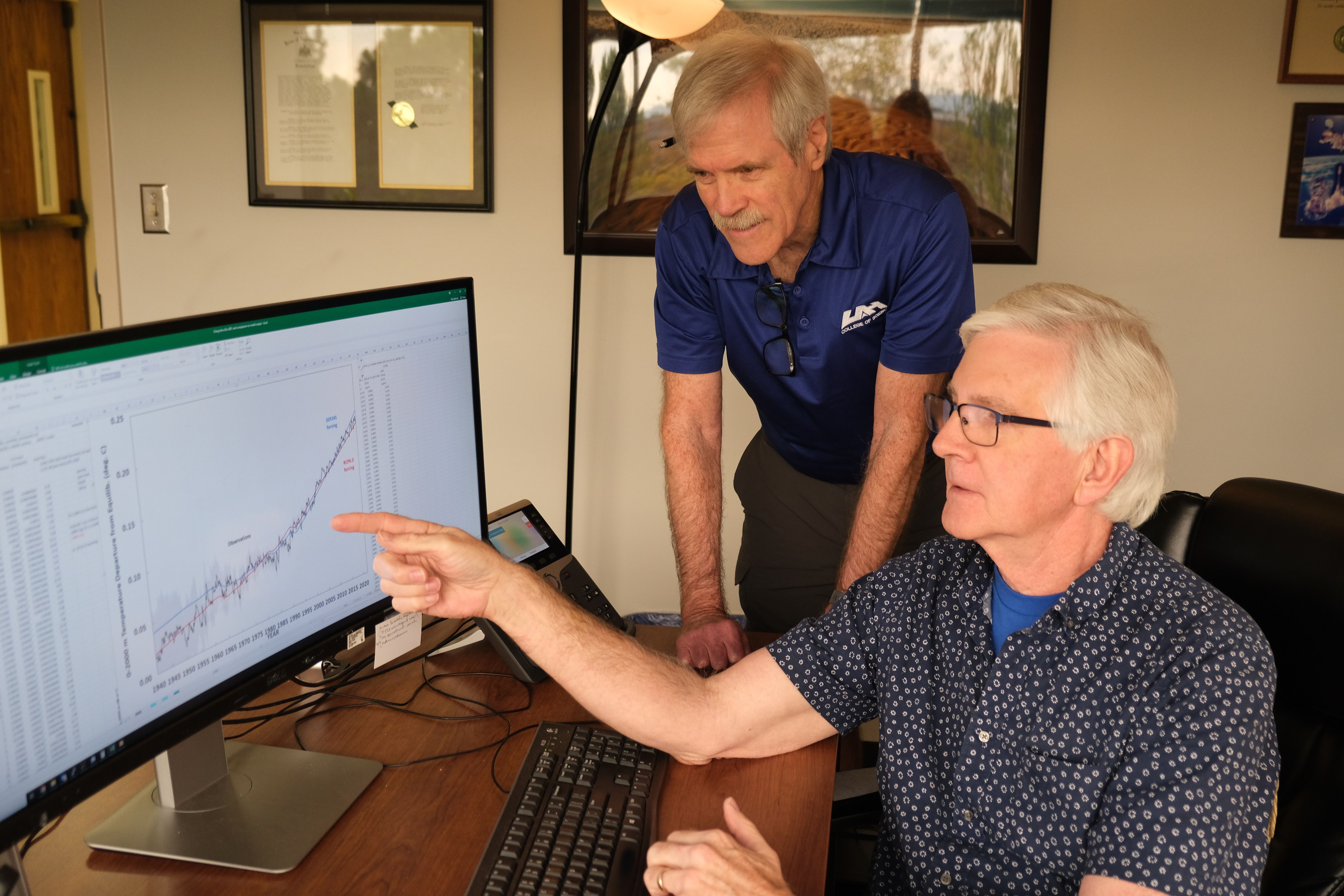 Two older men in an office examining a graph on a computer screen, one seated pointing at data, the other standing in a blue shirt with glasses hanging on his collar.