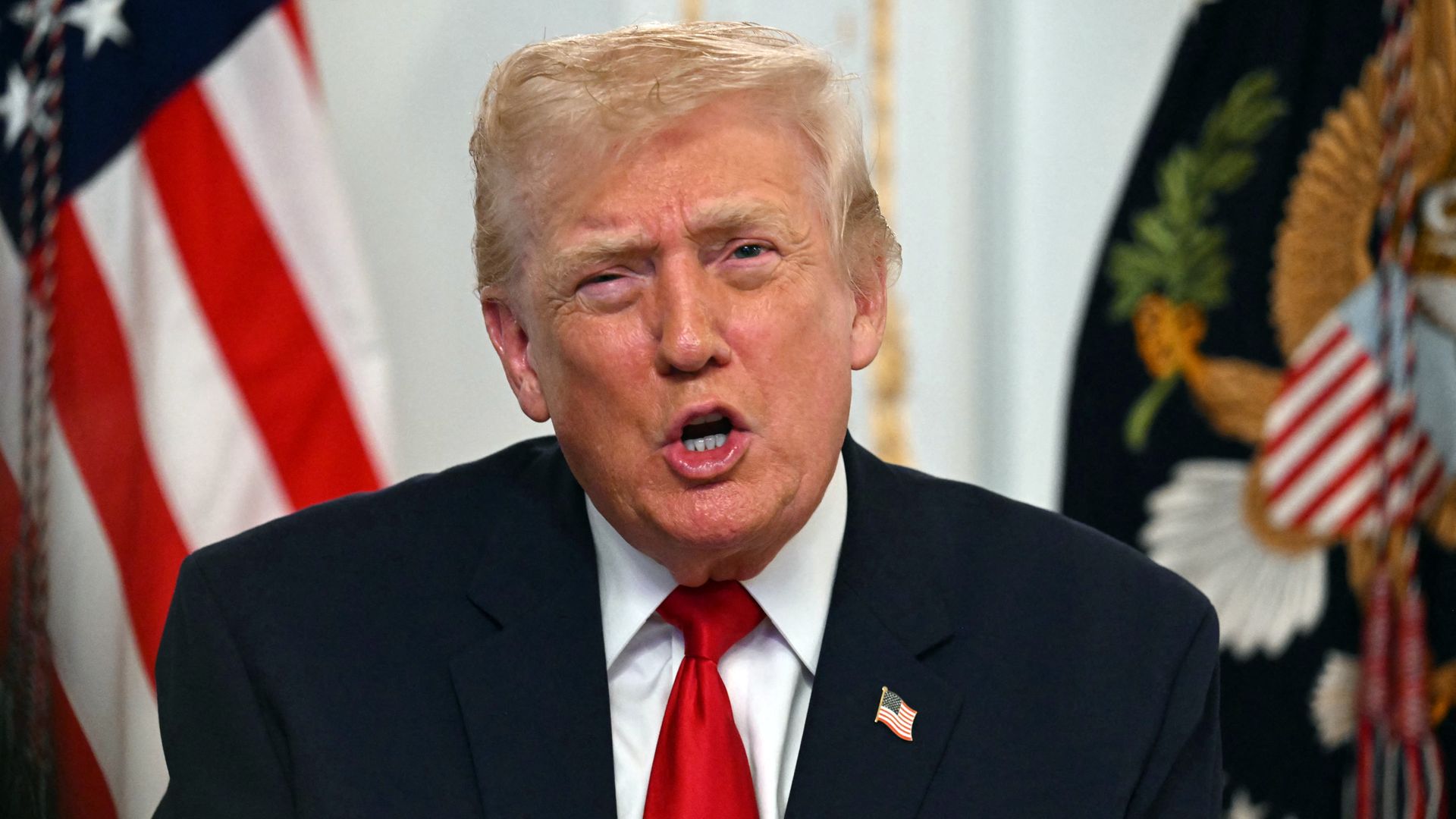 A gray-haired, fair-skinned President Trump, wearing a navy jacket with a US flag pin at the top of his left lapel, white shirt and red tie, talks in front of a US and another flag.
