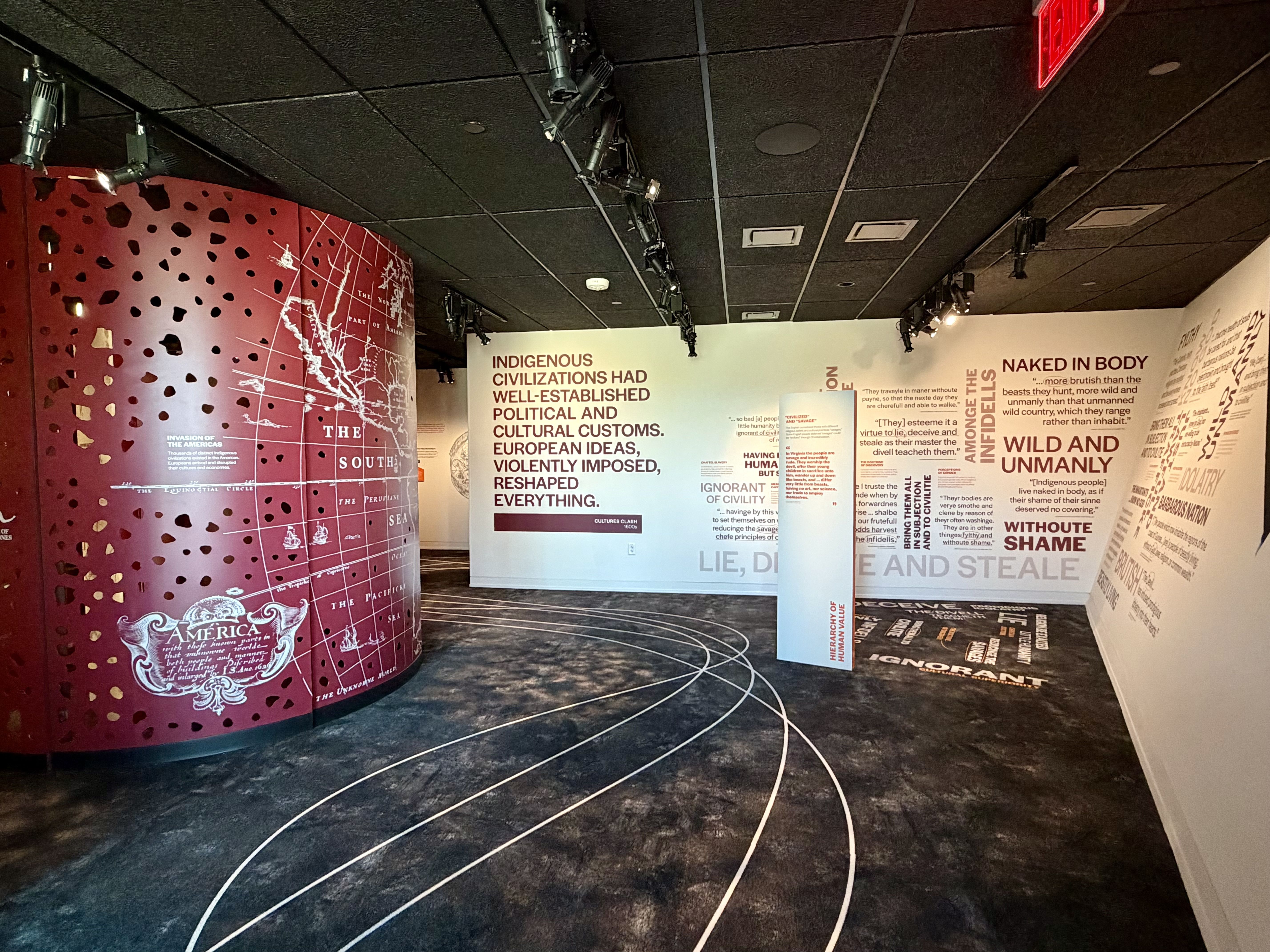 Exhibition gallery showing a maroon curved wall on the left with a perforated map design; white walls on the right feature bold red text panels and a central pillar, with curved floor lines.