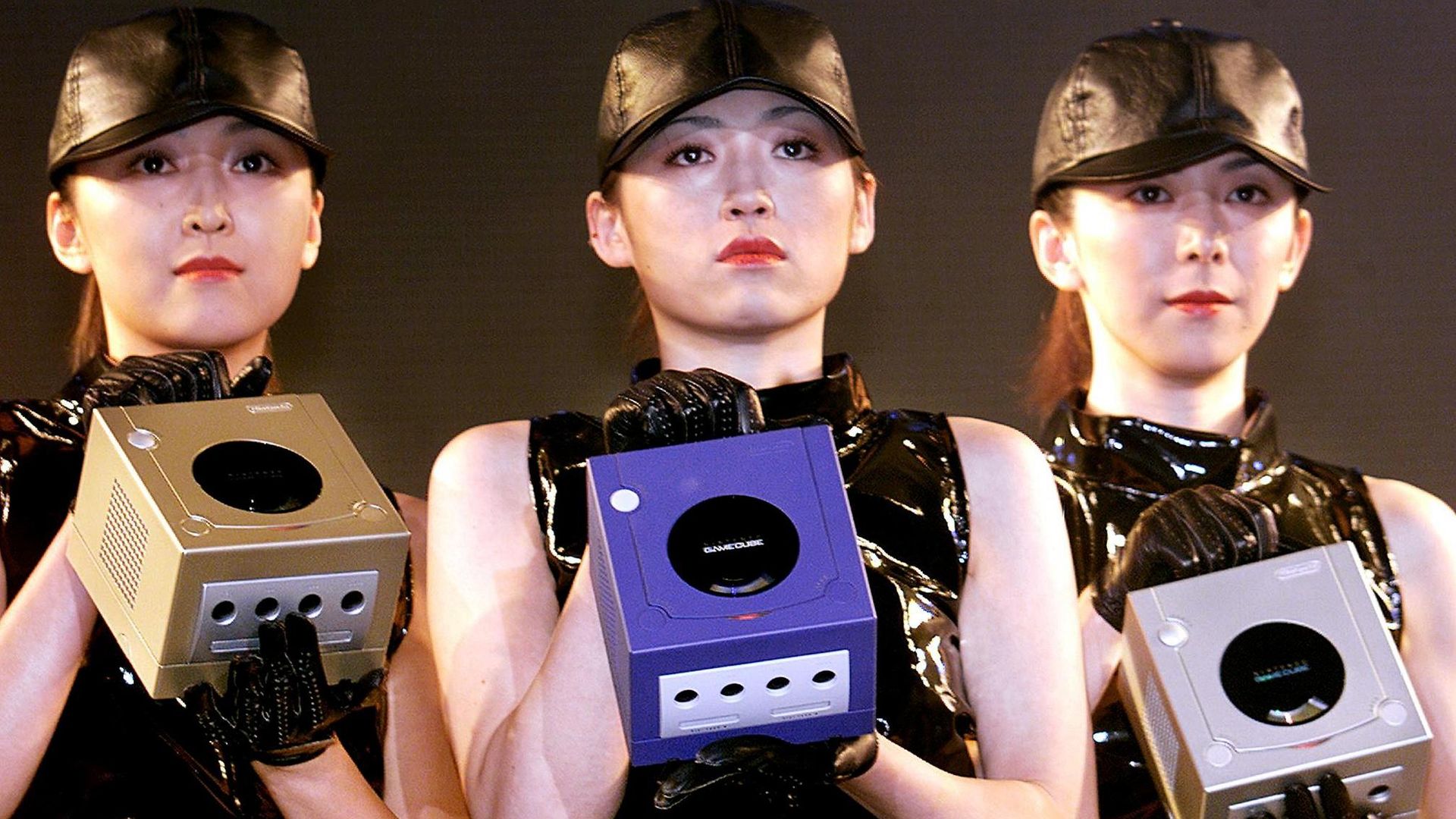 Three women in leather hats and tops holding cube-shaped gaming consoles.
