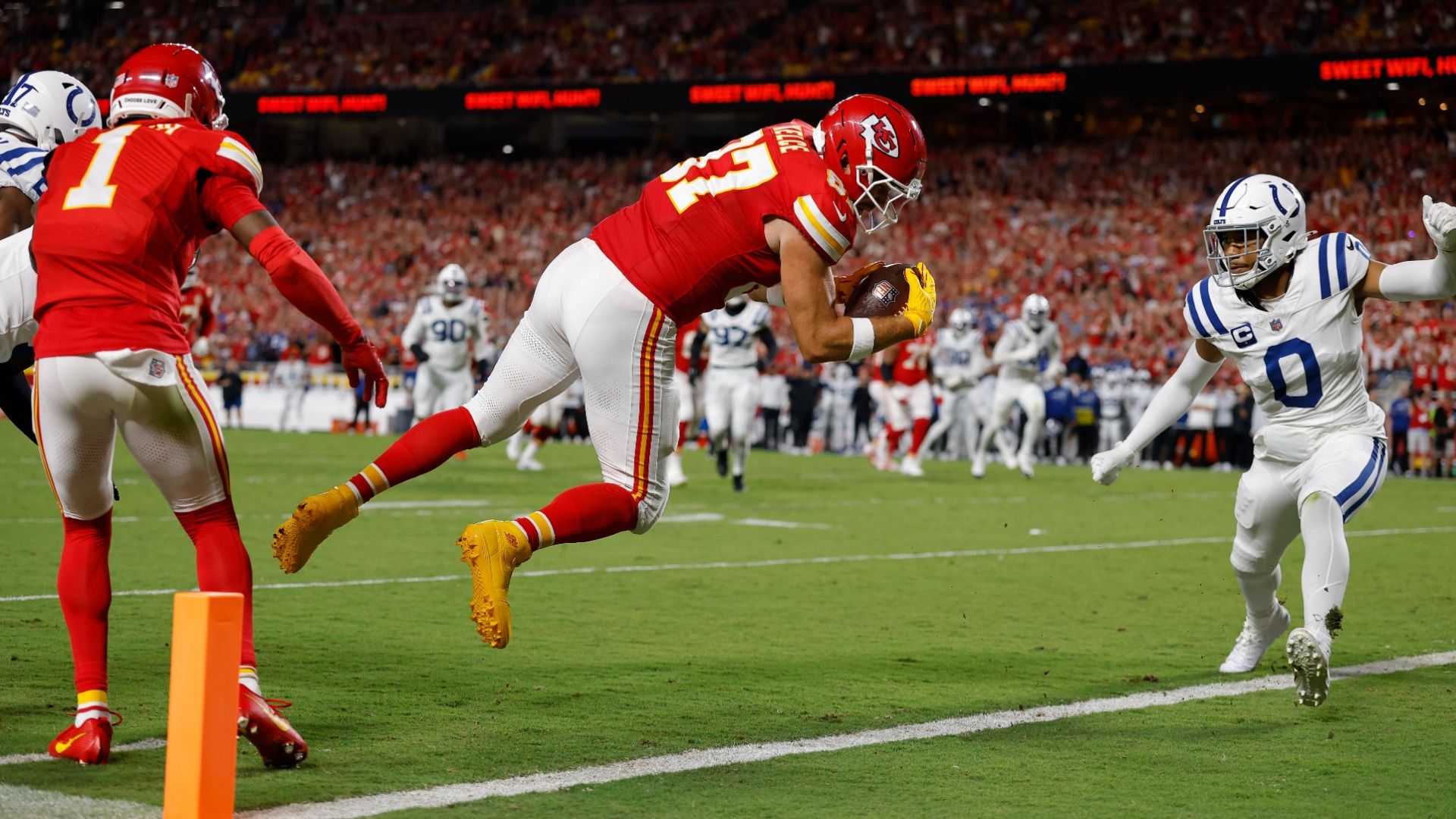 Chiefs player No. 87 Travis Kelce dives midair over the goal line while Colts No. 0 runs up to try and stop him, with an orange pylon in the foreground and other players and a crowd in the background.