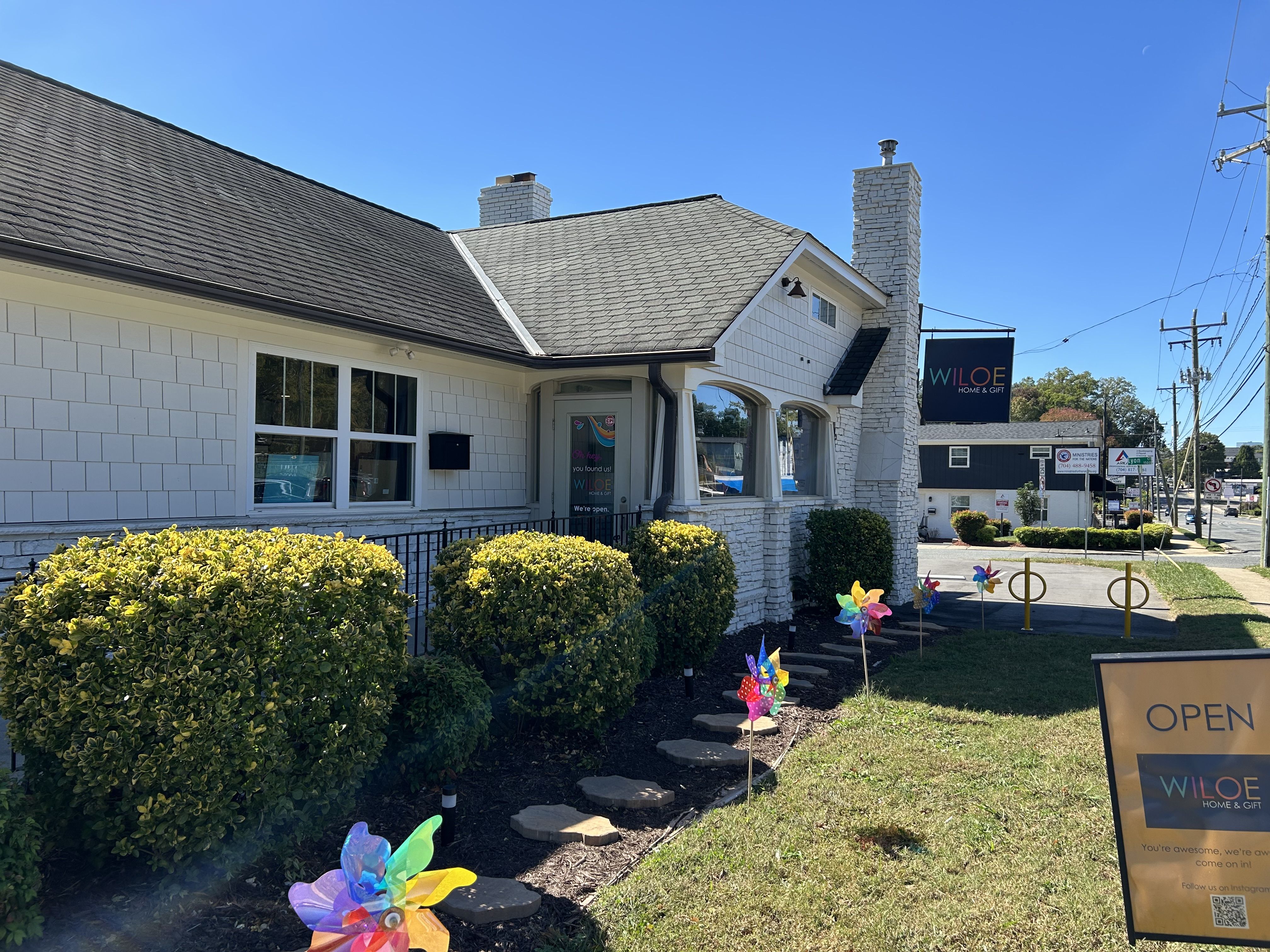 White building with black roof and stone chimney, green shrubs, colorful pinwheels along stone path, sign reading "OPEN WILOE Home & Gift" under clear blue sky.
