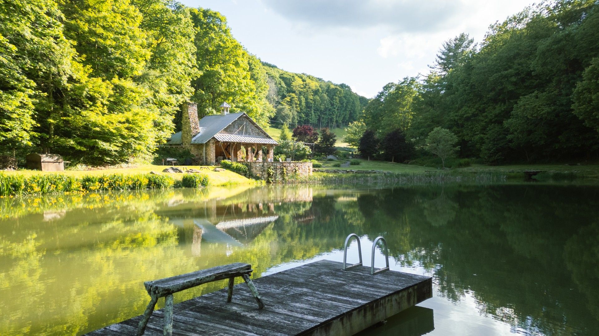 A rustic stone house with a covered porch overlooks a peaceful pond with a wooden dock and lush greenery.