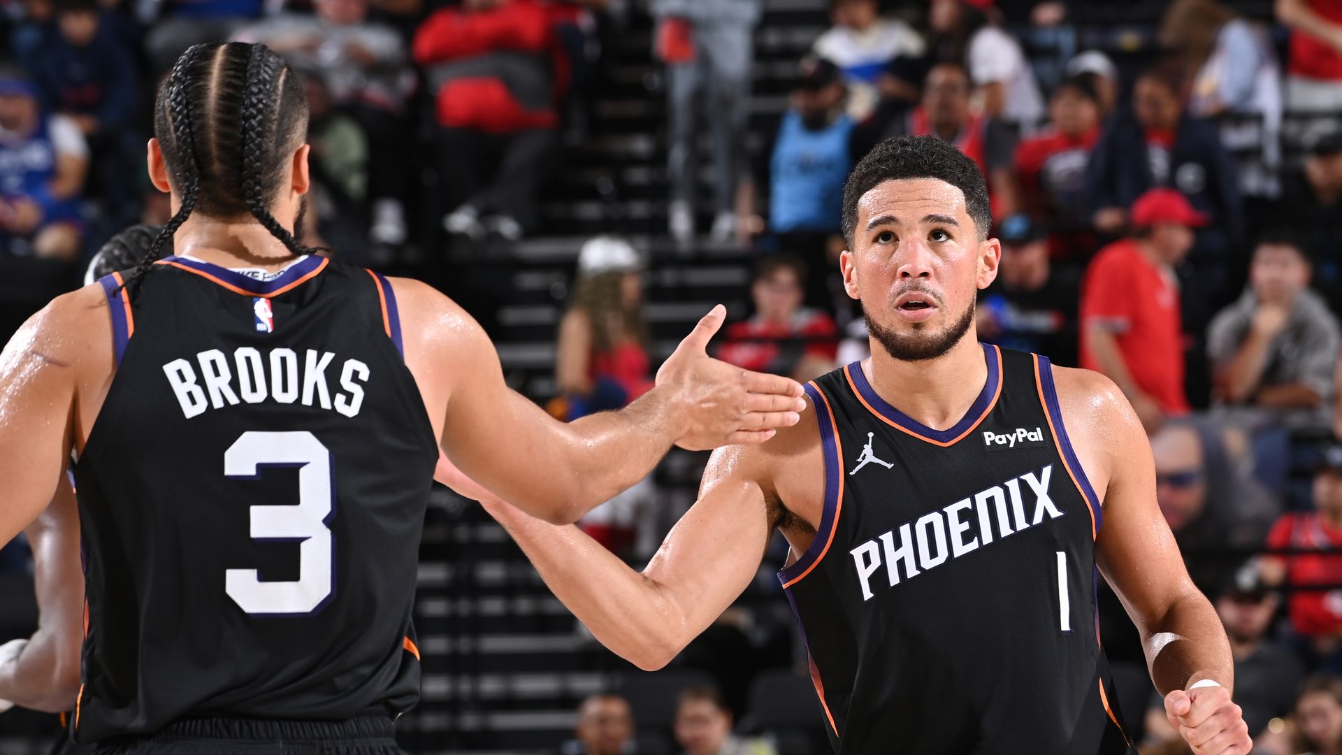 Two basketball players in black uniforms with white lettering, one facing the camera and one with his back to the camera, slap hands on the court. 