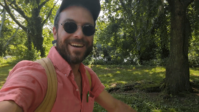 A man wearing a pink shirt and dark-blue hat moving quickly in a wooded area with a river in the background.