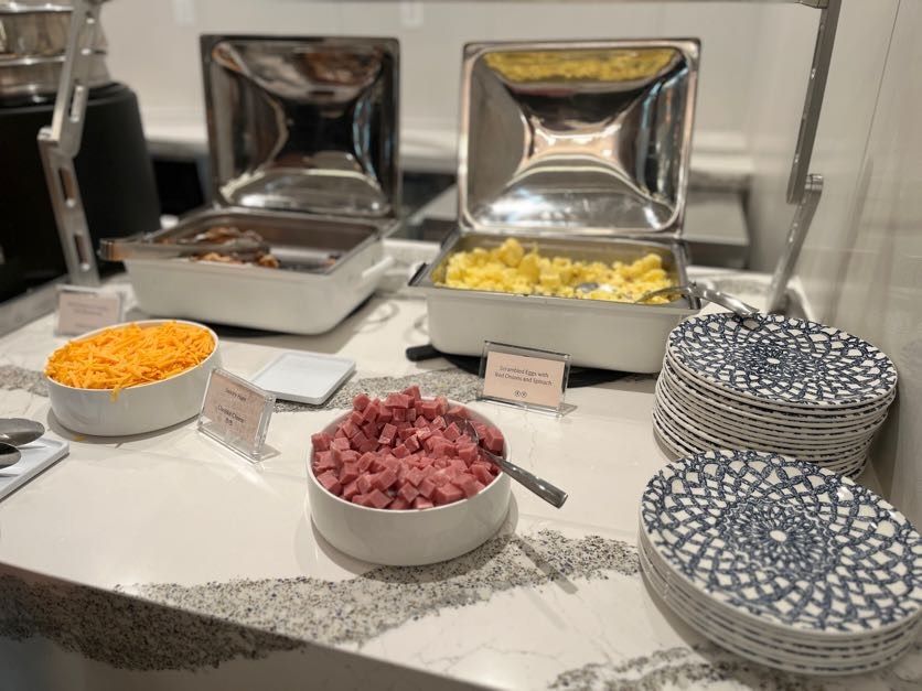 Small white bowls of shredded cheese and ham in front of stainless steel trays of scrambled eggs with blue and white plates on the side.