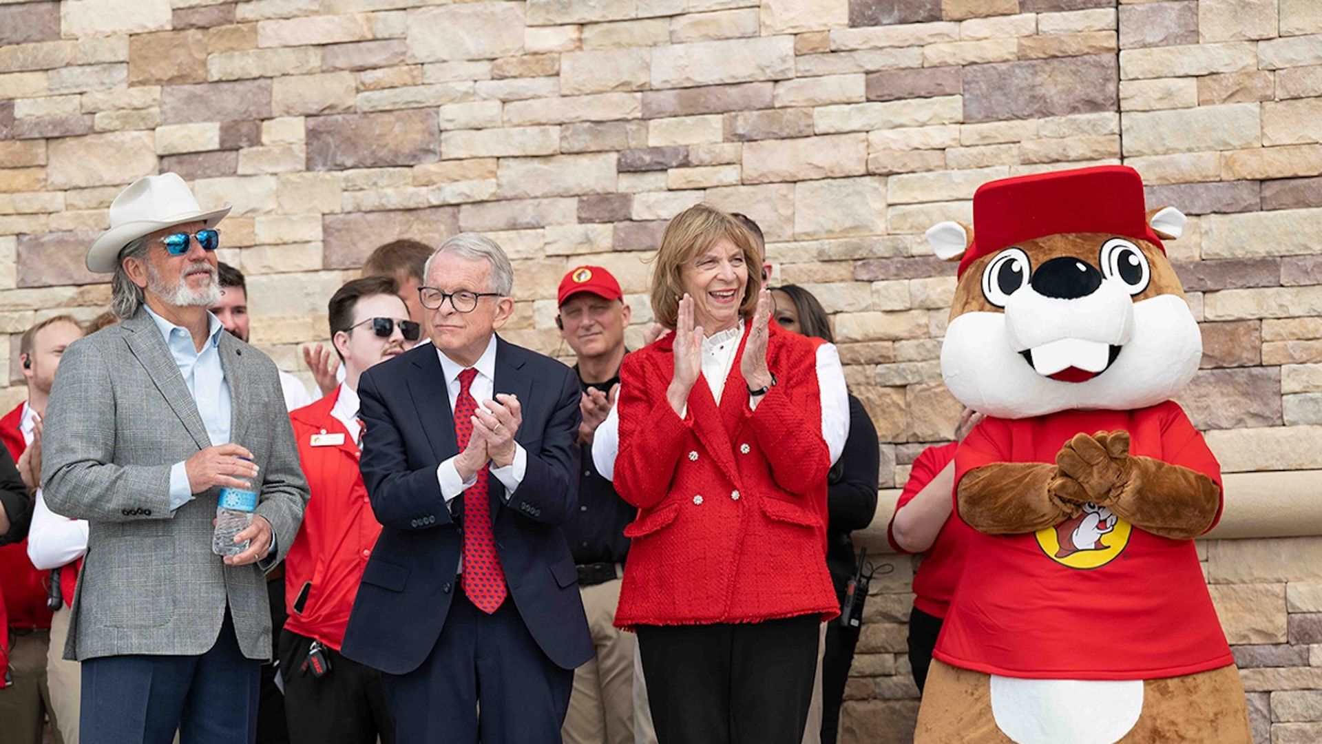 Group photo in front of a beige stone wall: man in gray suit and white cowboy hat stands next to Ohio Gov. Mike DeWine (dark suit with red tie), DeWine's smiling wife Fran in a red jacket, and a large Buc-ee's beaver mascot in a red shirt; others in red behind.