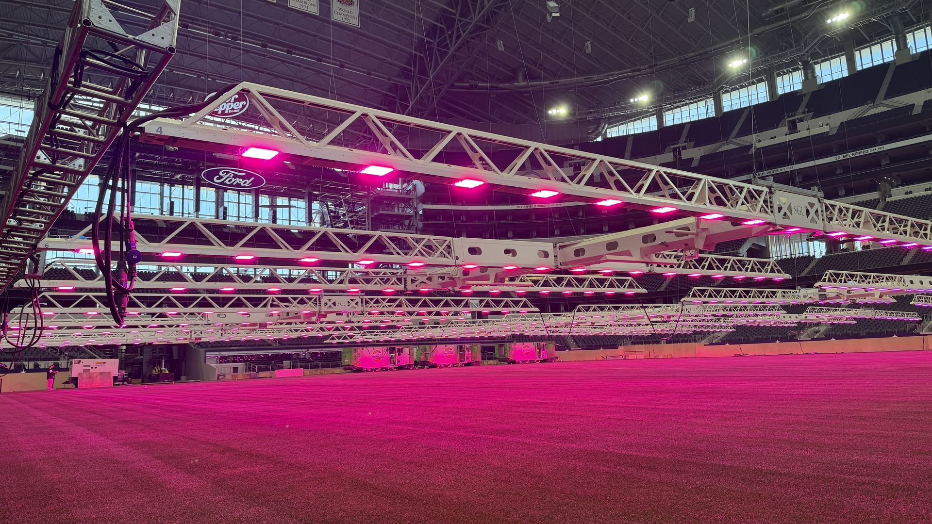 Inside a large arena with white truss structures overhead, pink lights line the beams. A Ford logo sign is visible among the rigging; empty seating and a pink-colored floor.