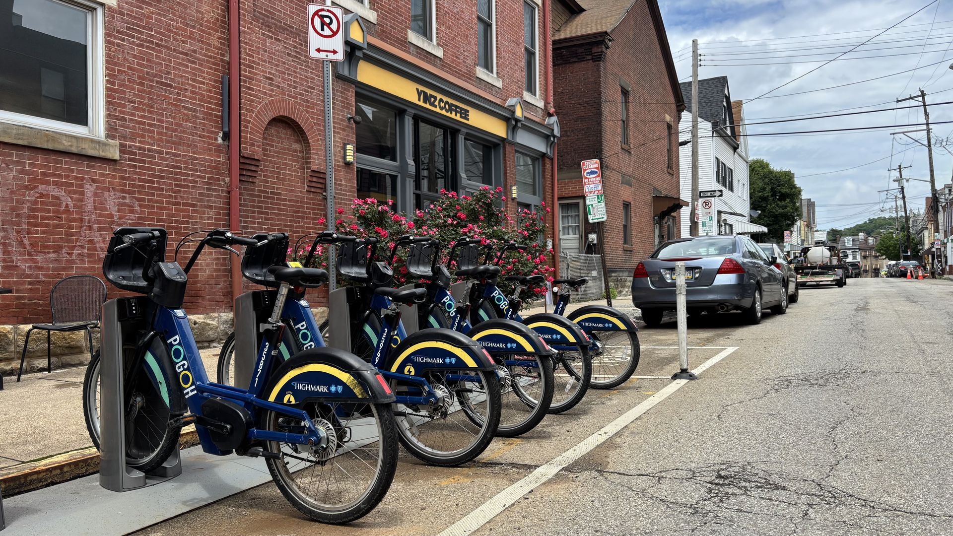 Street scene of red brick buildings, a bike-share dock in the foreground, pink flowers, a yellow "YIZ COFFEE" sign, a parked car, no-parking sign, and overhead power lines.