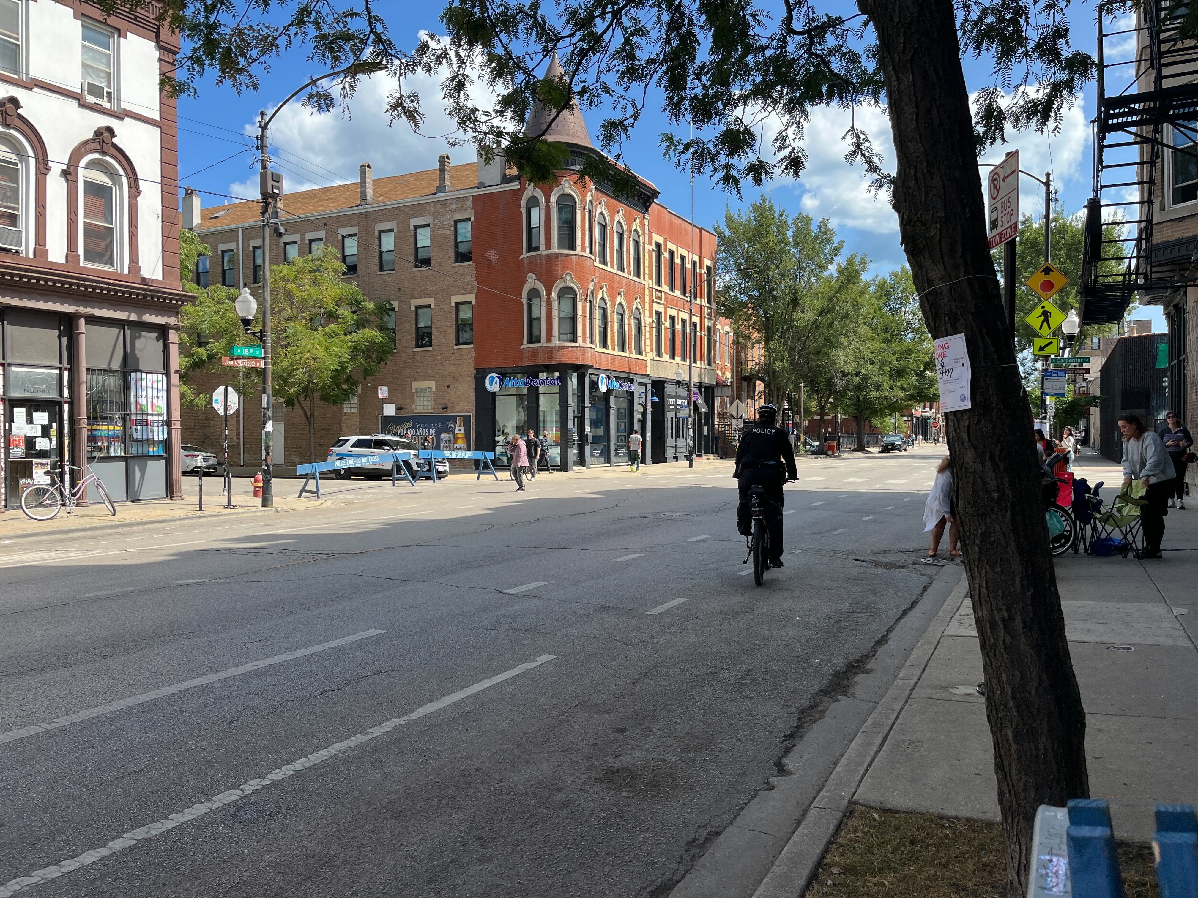 City street scene with a red brick corner building housing "Alto Dental," a police officer riding a bike, pedestrians on sidewalk, a police car, and trees under a partly cloudy blue sky.