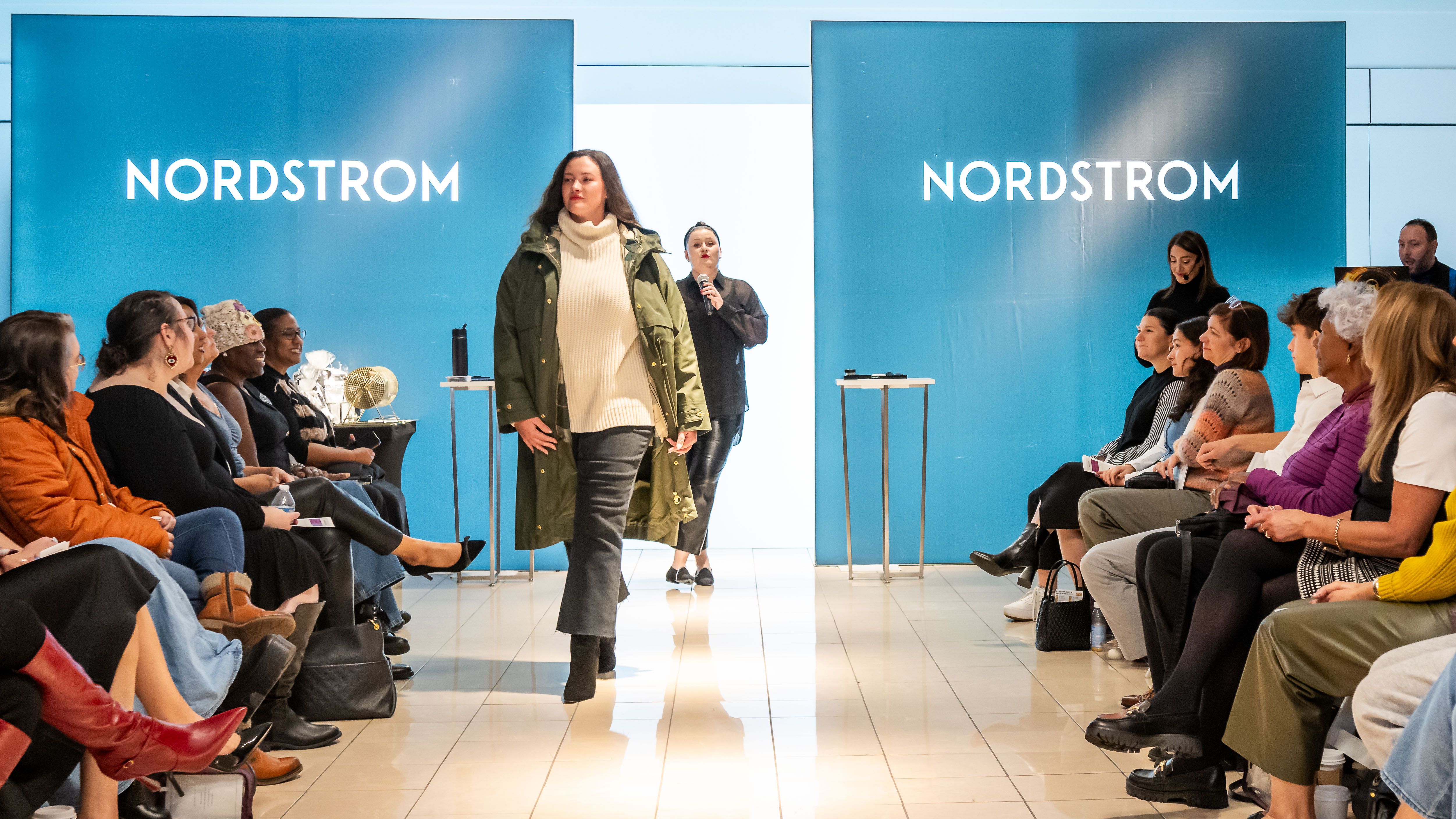 Fashion show at Nordstrom with a model walking on runway in green jacket and white sweater, audience seated on both sides watching attentively.