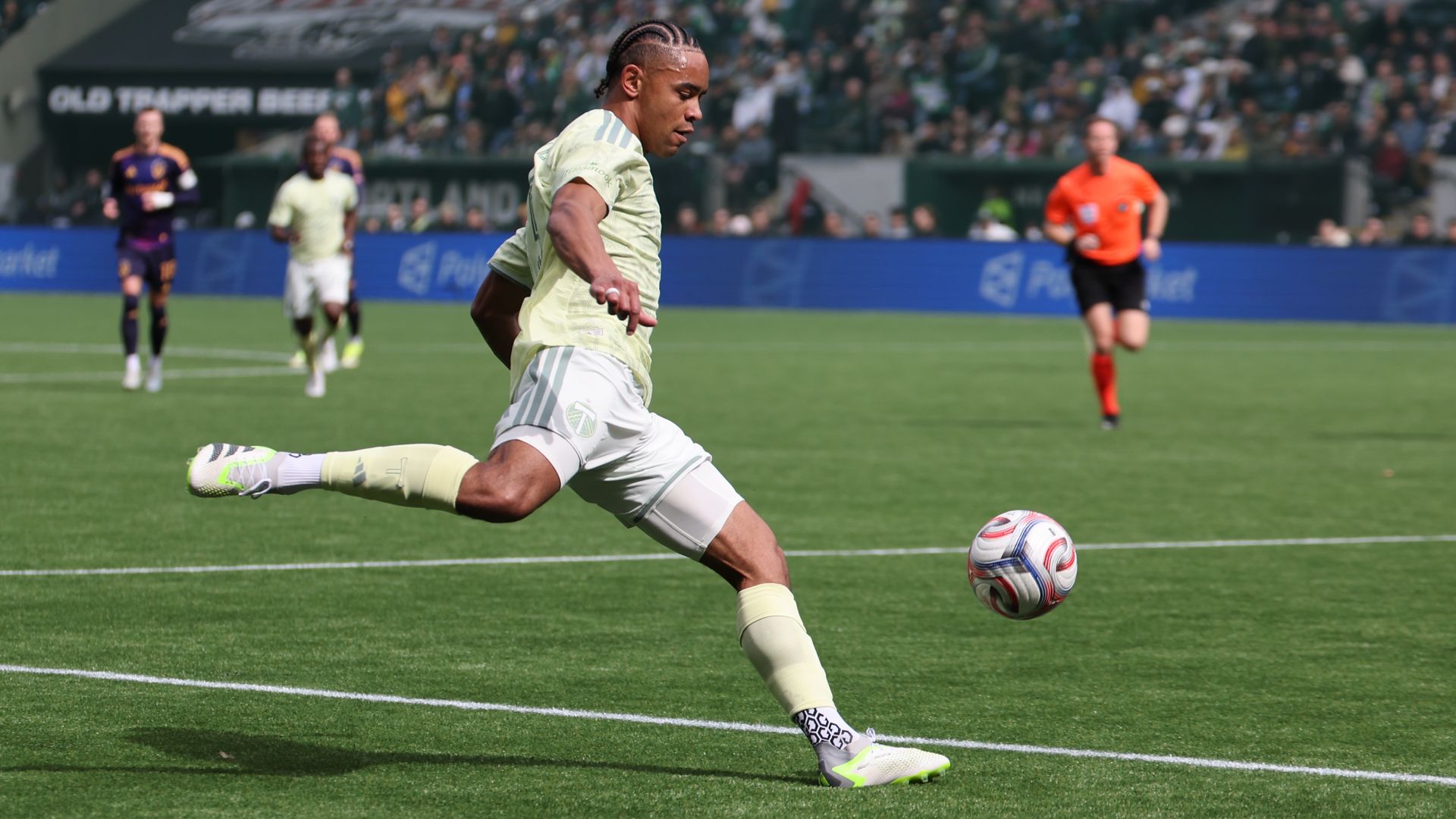 Soccer player in a pale yellow kit kicks a white ball on a green pitch, with teammates, an orange referee, and a crowded stadium in the background watching the action.
