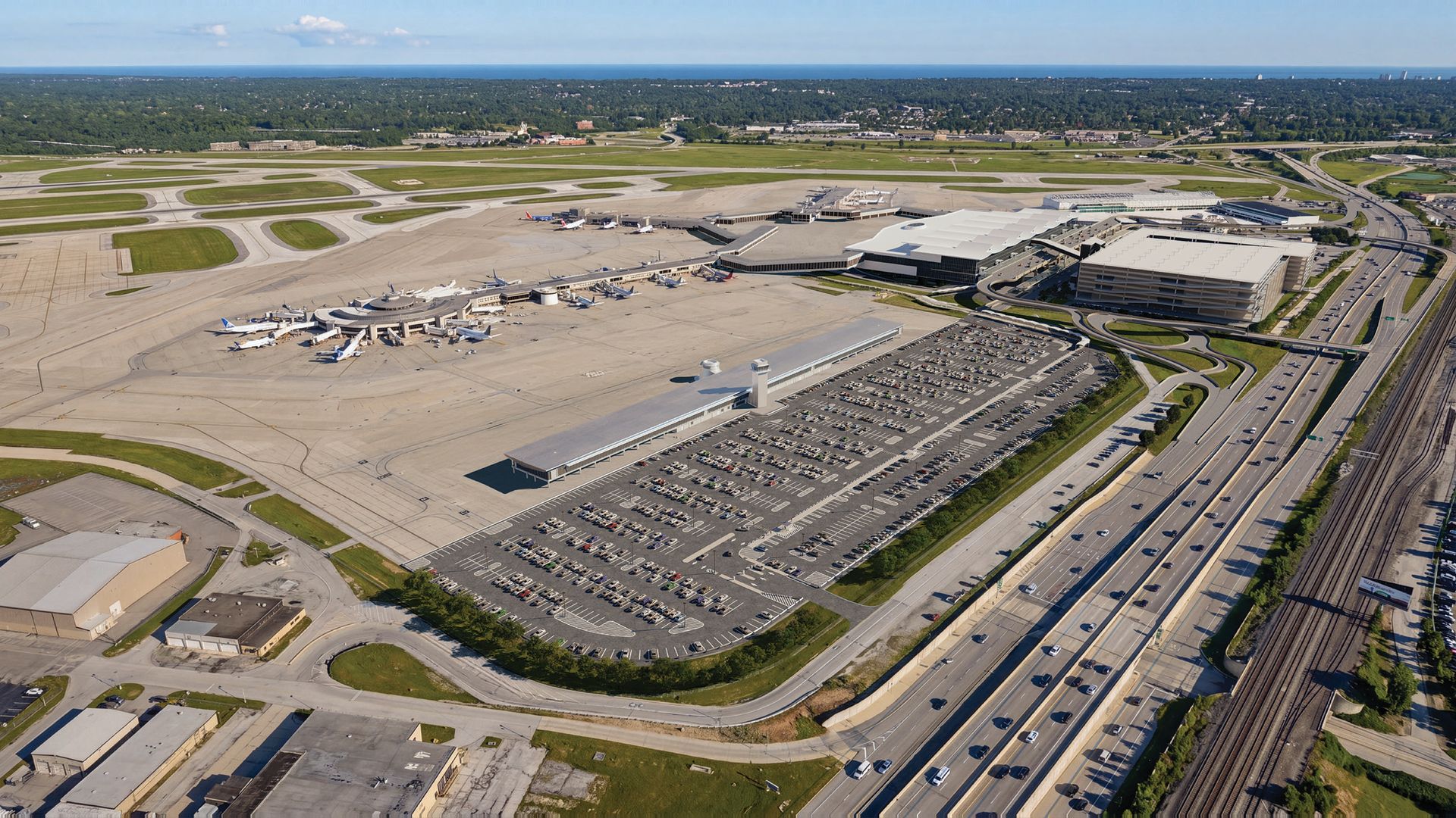 An aerial view of Cleveland Hopkins International Airport: a large terminal with gates and planes, extensive tarmac, taxiways, adjacent parking lot, and surrounding highways and green fields.