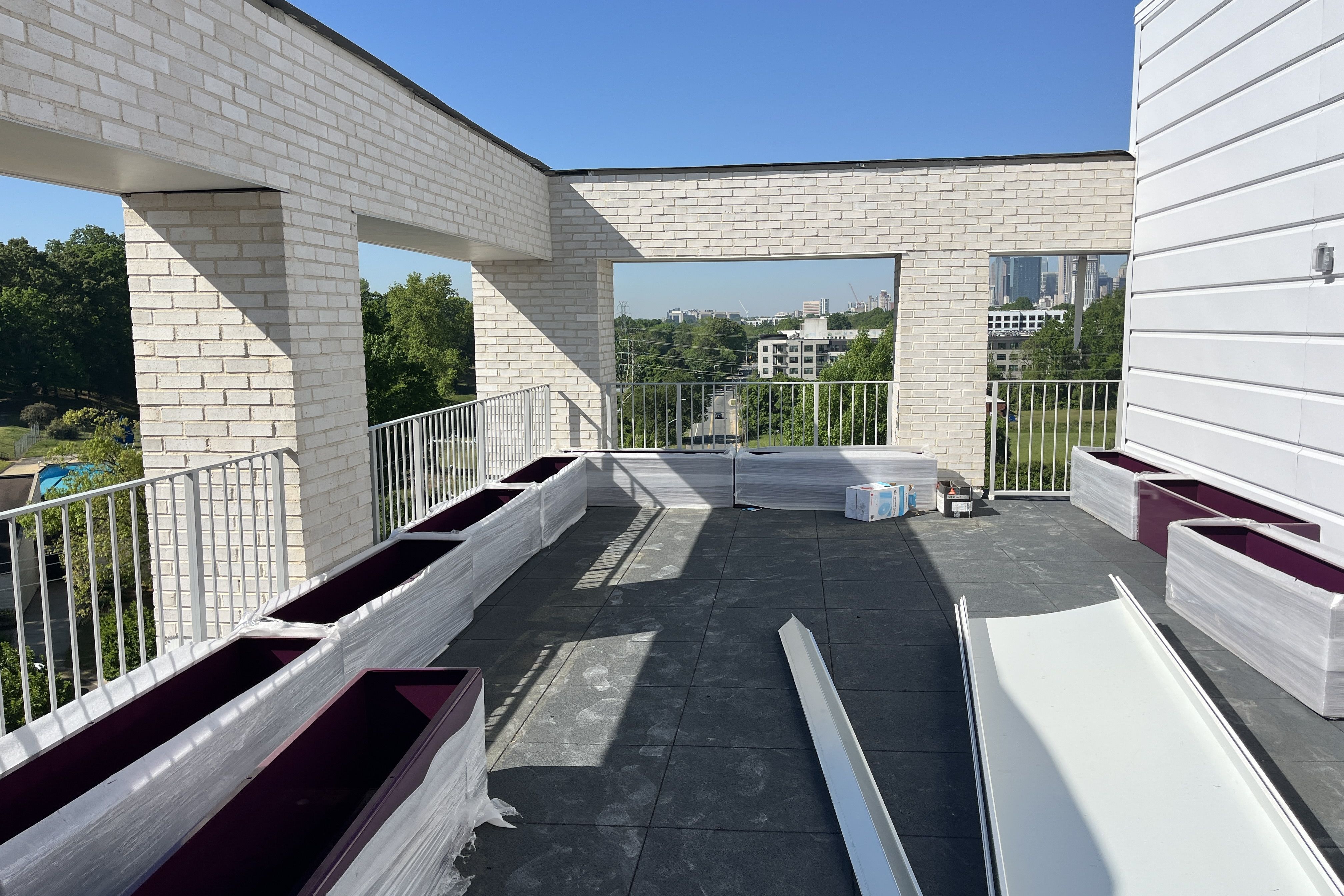 Rooftop scene of a white brick structure with window openings, gray decking, and white railings. Construction materials lie nearby; city skyline visible under a clear blue sky.