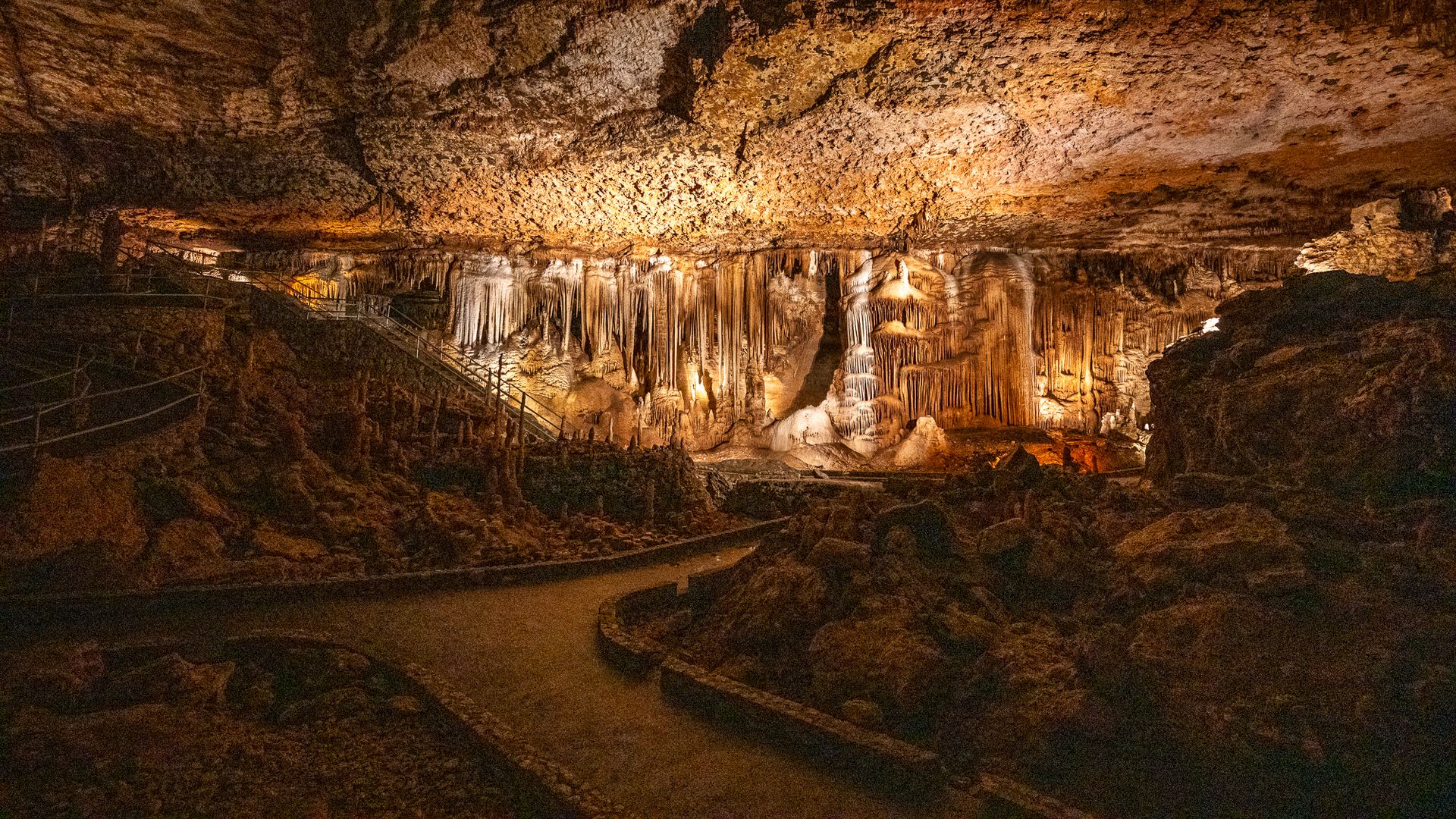 An illuminated underground cavern with dramatic stalactites and stalagmites, featuring a winding path and rock formations.