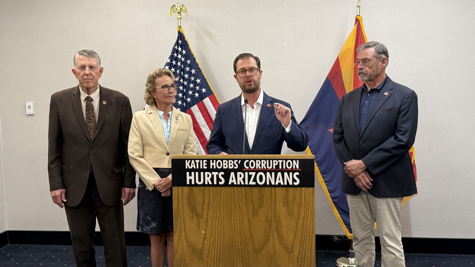 A man in a blue suit jacket and white shirt gestures with his left hand in front of him while he speaks at a microphone behind a lectern with a sign reading, "KATIE HOBBS' CORRUPTION HURTS ARIZONANS"; US and Arizona flags are against white wall behind him, and three people stand to his sides.