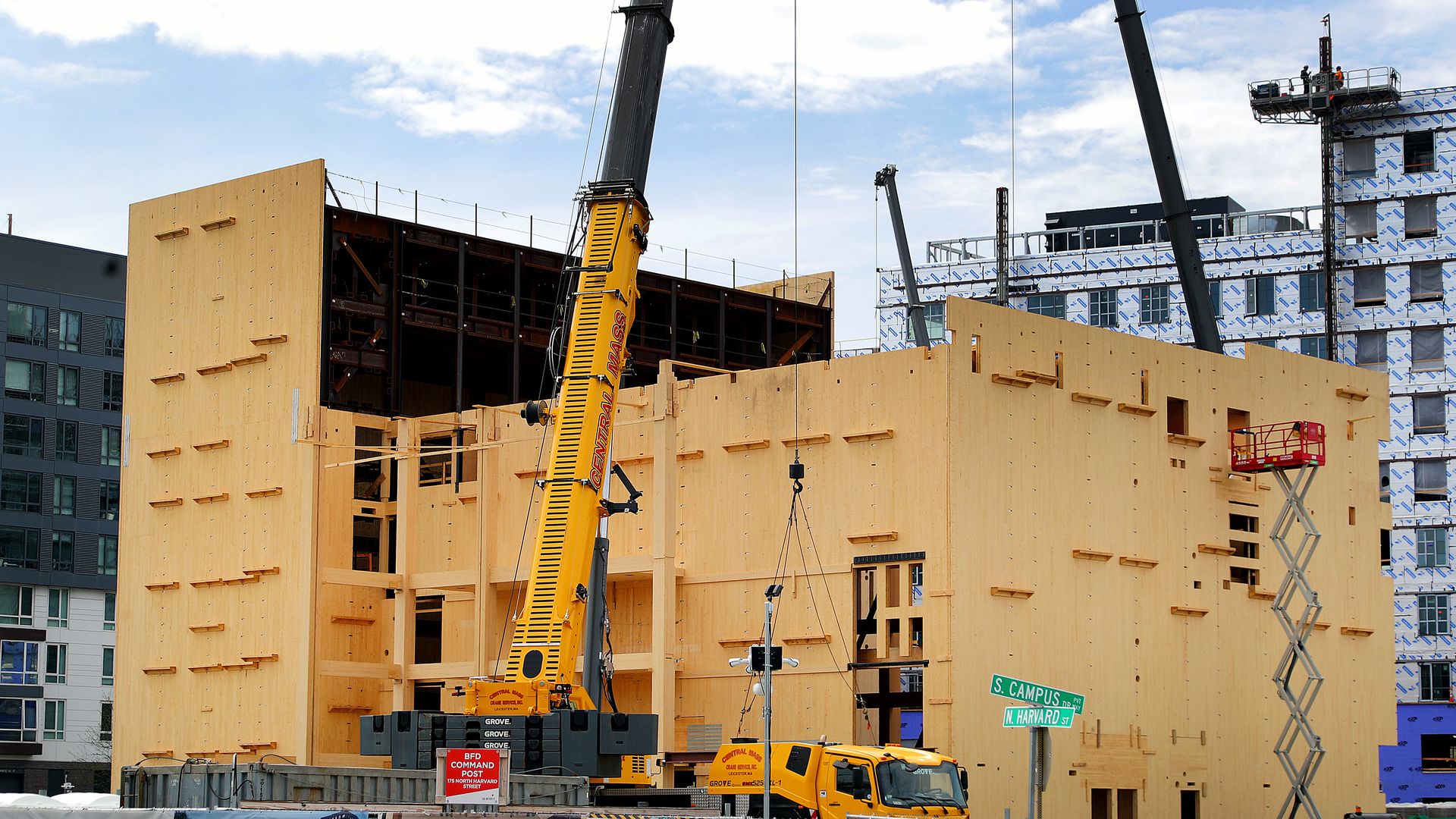 Construction site with yellow cranes and a large wooden building frame under cloudy sky. Signs indicate "DO NOT ENTER" and "SIDEWALK CLOSED" near South Campus and North Harvard streets.