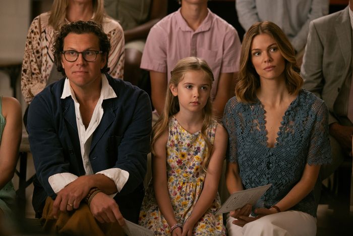 A man in glasses and navy jacket, a young girl in a floral dress, and Brooklyn Decker in a blue lace top sit closely together, appearing attentive in an indoor setting with others behind.
