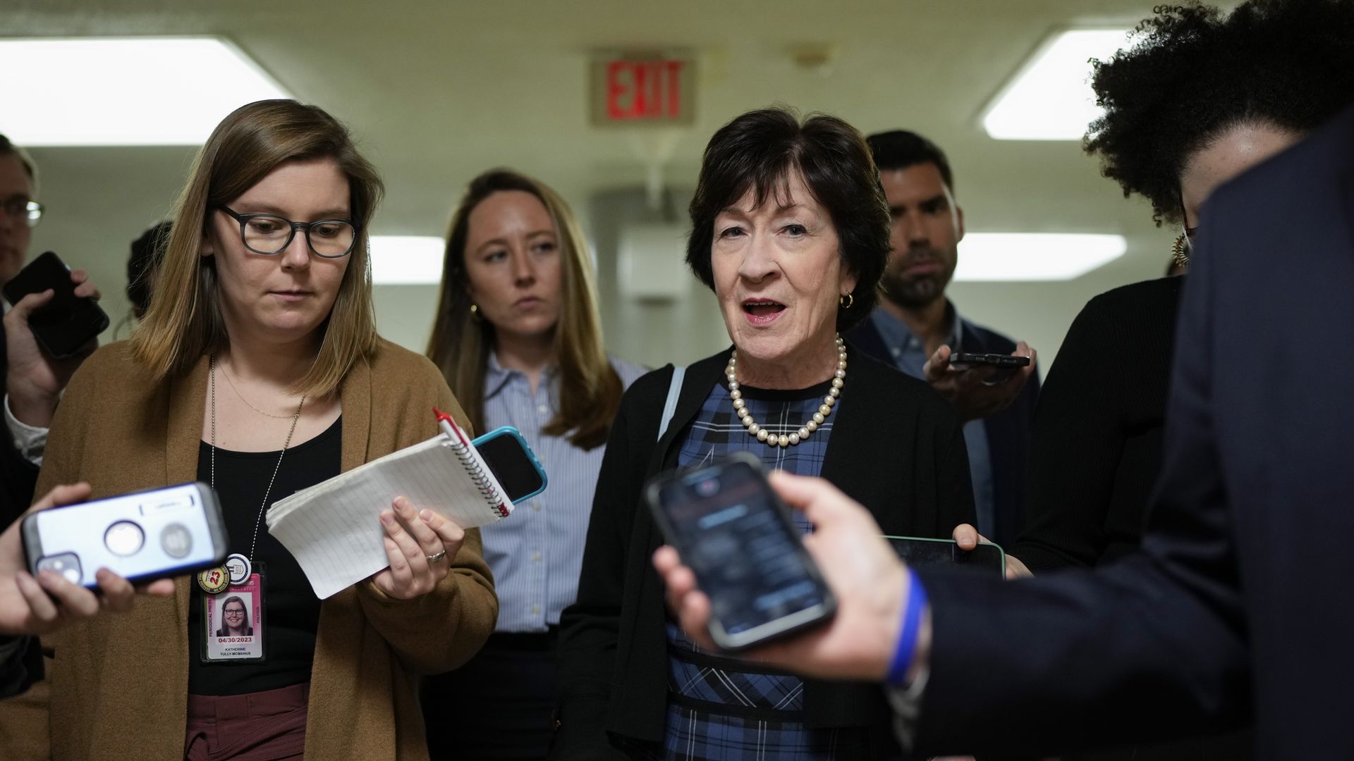 Sen. Susan Collins, wearing a black jacket, blue plaid dress and pearl necklace, speaks to reporters in the Capitol basement.