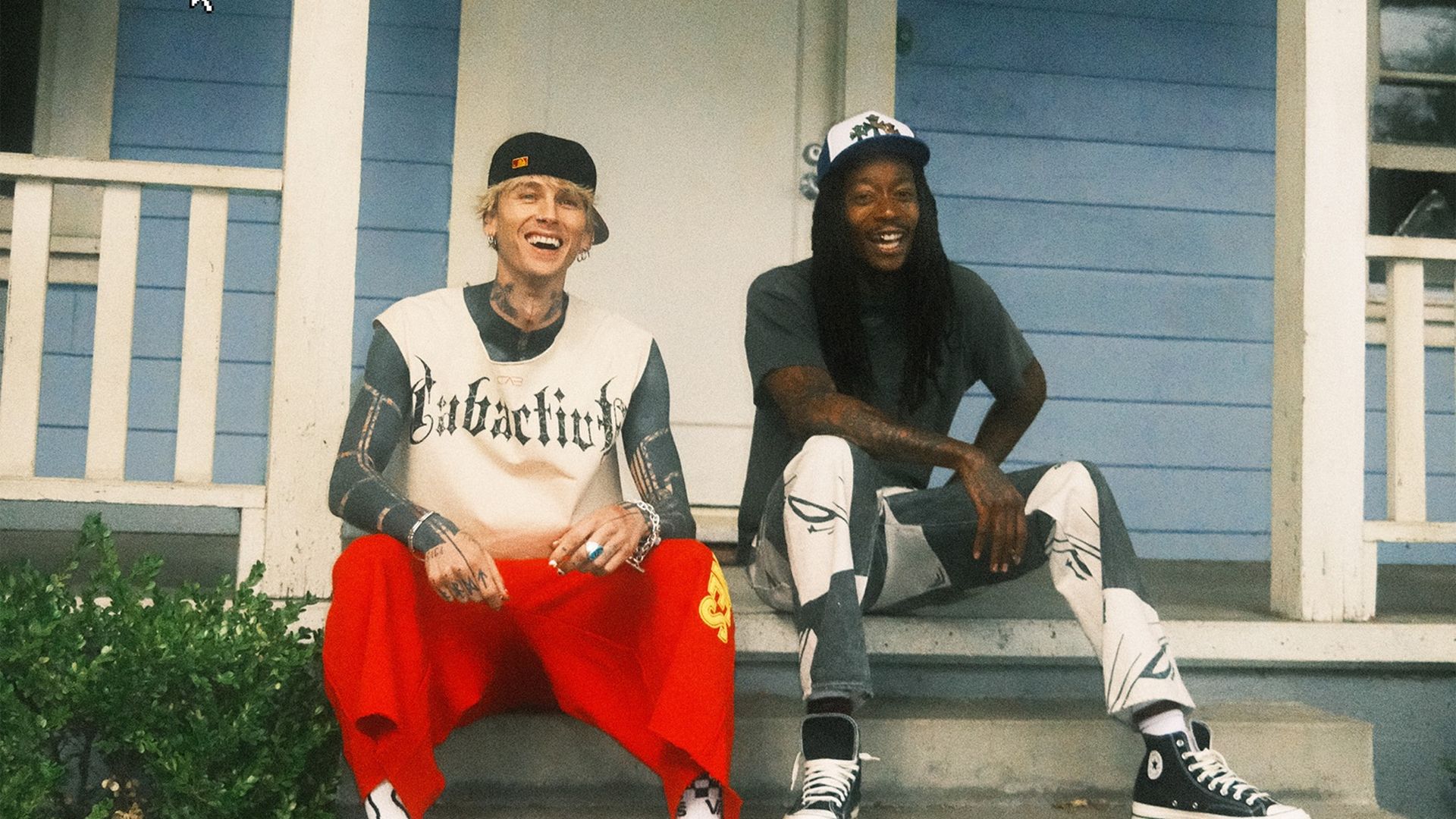 Two friends sit on the blue front porch steps of a house, smiling. Left: blond with backward black cap, white graphic tee, bright red pants. Right: dark-haired with white cap and gray shirt.