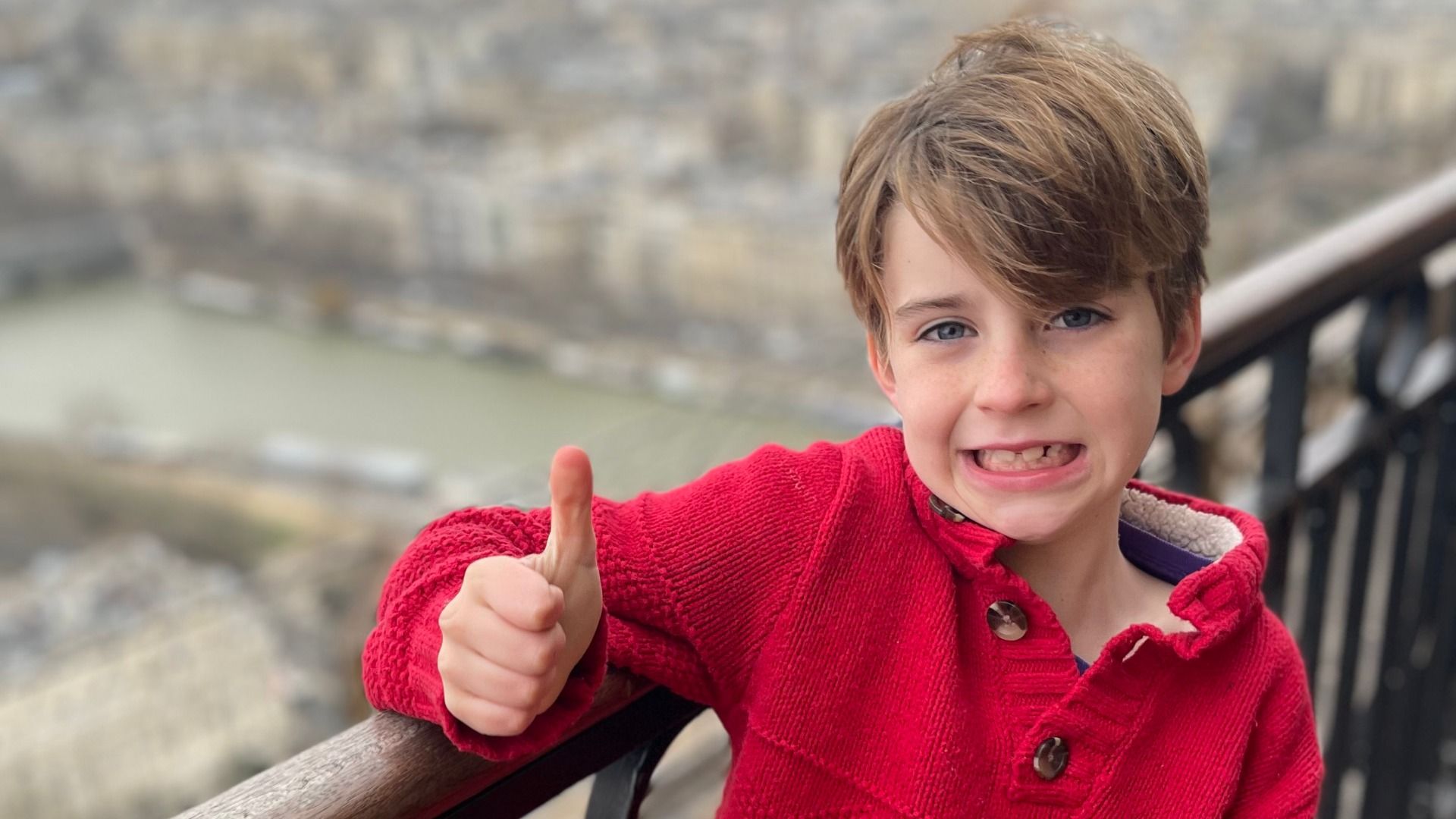Young boy with light brown hair wearing a red sweater gives a thumbs-up while standing by a railing overlooking a blurry cityscape with a river in the background.
