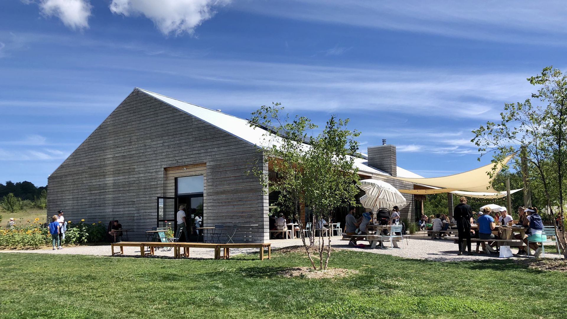 The brewery building is pictured open with blue sky and green grass and bustling with customers.