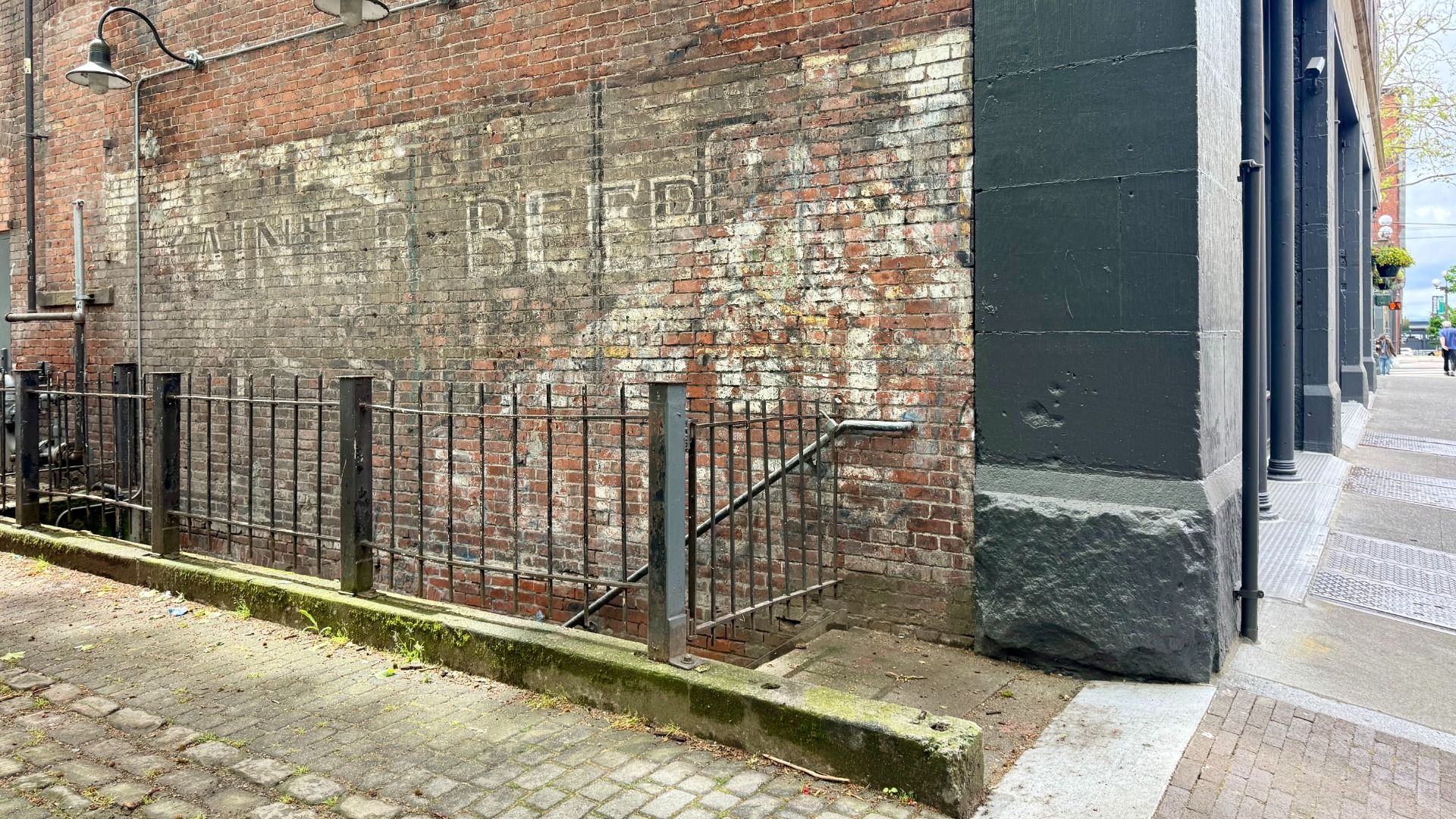Wide urban scene: a weathered brick wall with a faded painted sign, a rusty iron railing in front, and the dark stone corner of a building on the right; sidewalk and distant pedestrians.