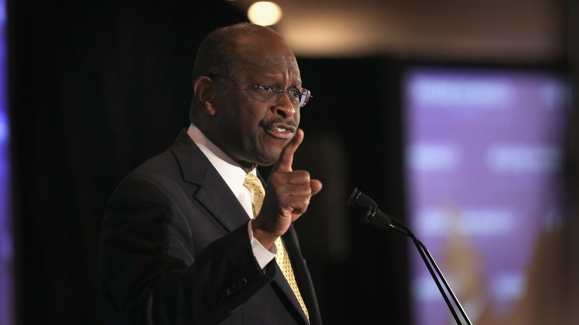 In this image, Herman Cain points a finger in the air while speaking into a microphone. 
