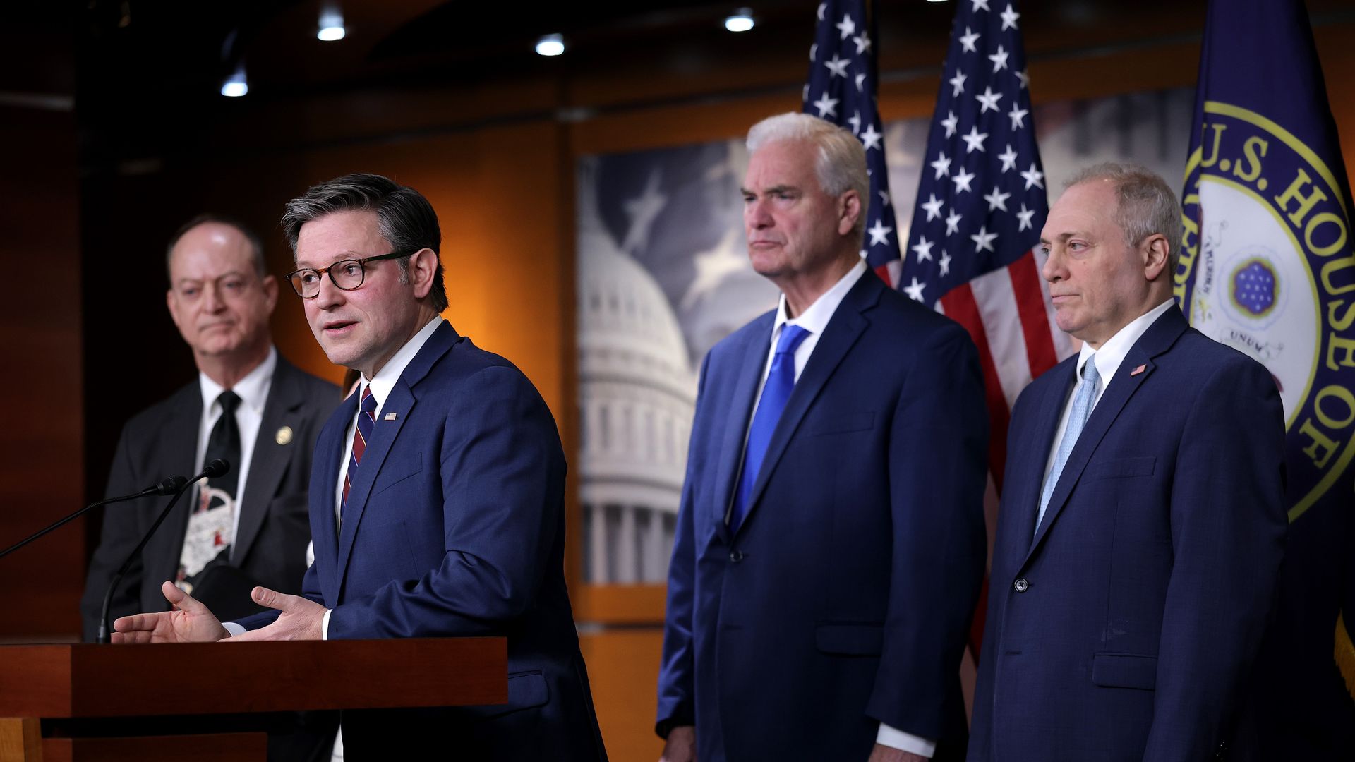 Four men in suits stand at a podium with U.S. flags and a U.S. House of Representatives seal in the background; the man at the podium speaks while others listen attentively.