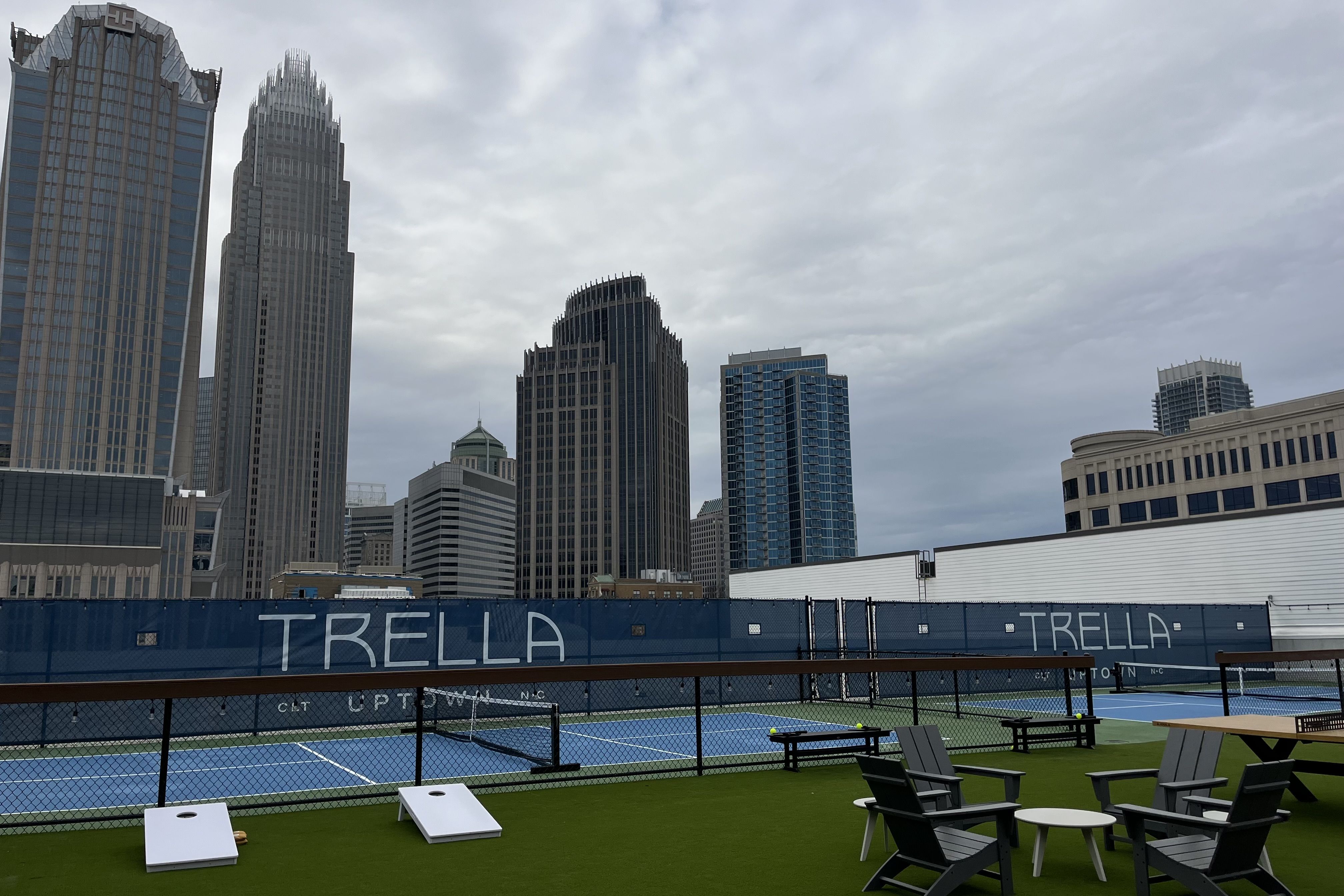 Outdoor tennis court with blue courts and green artificial turf, surrounded by black fence and seating area with Adirondack chairs, under cloudy sky and tall city buildings in background.