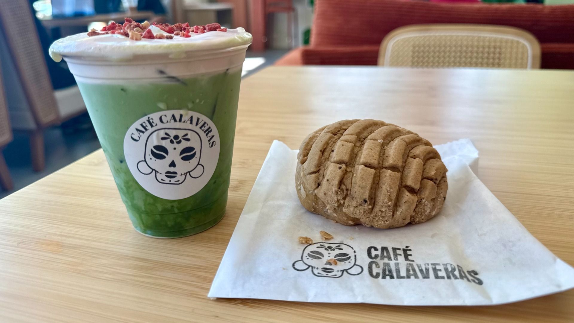 Iced green matcha drink with cream and red toppings in a Café Calaveras cup, next to a brown concha pastry on a Café Calaveras napkin, on a wooden table in a café.