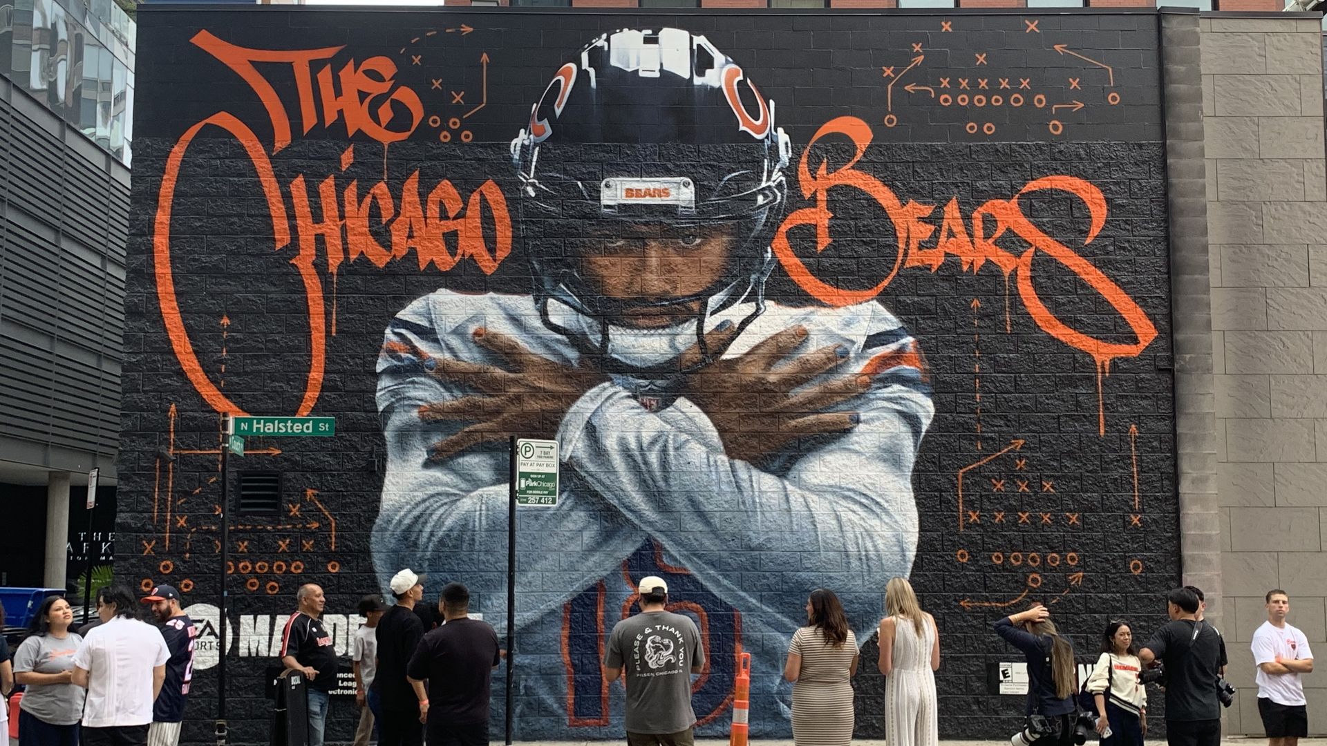 Mural on a black brick wall features a person in a white astronaut suit with an orange helmet, orange graffiti text and circuitry patterns; street sign shows N Halsted St, and people stand in front.