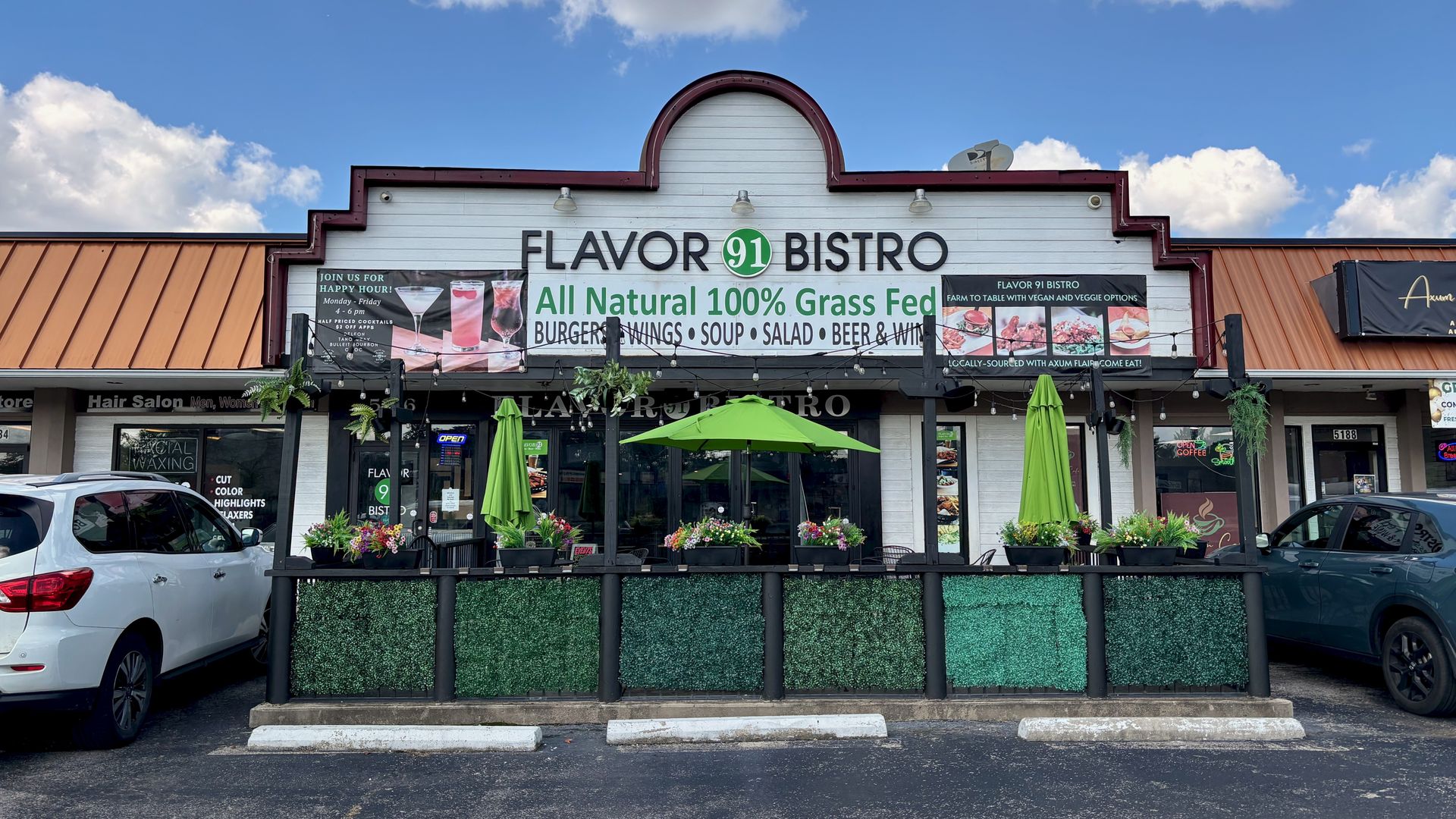 Exterior of Flavor 91 Bistro with white sign, green umbrella, green umbrellas on patio, and greenery under a partly cloudy sky.