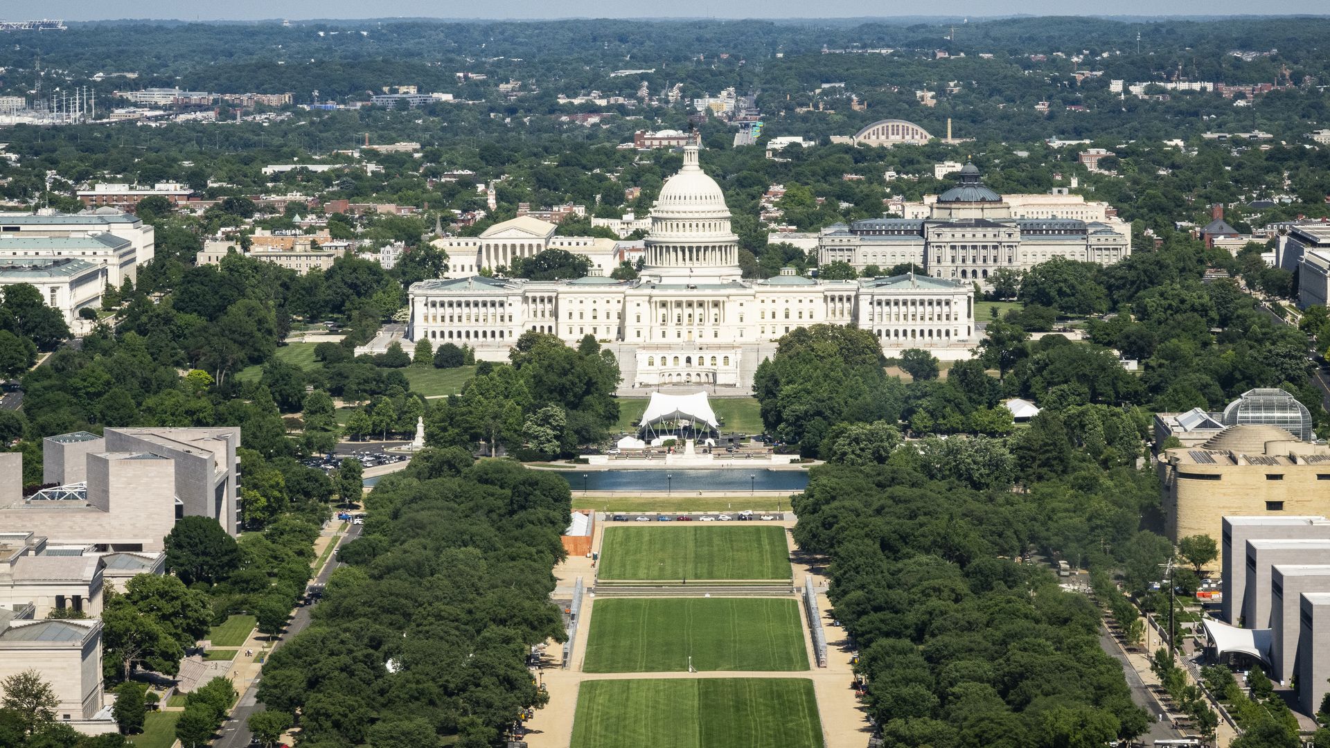 Aerial view of the U.S. Capitol with its domed rotunda, flanked by buildings and green lawns, dense trees, and a distant cityscape under a clear blue sky.