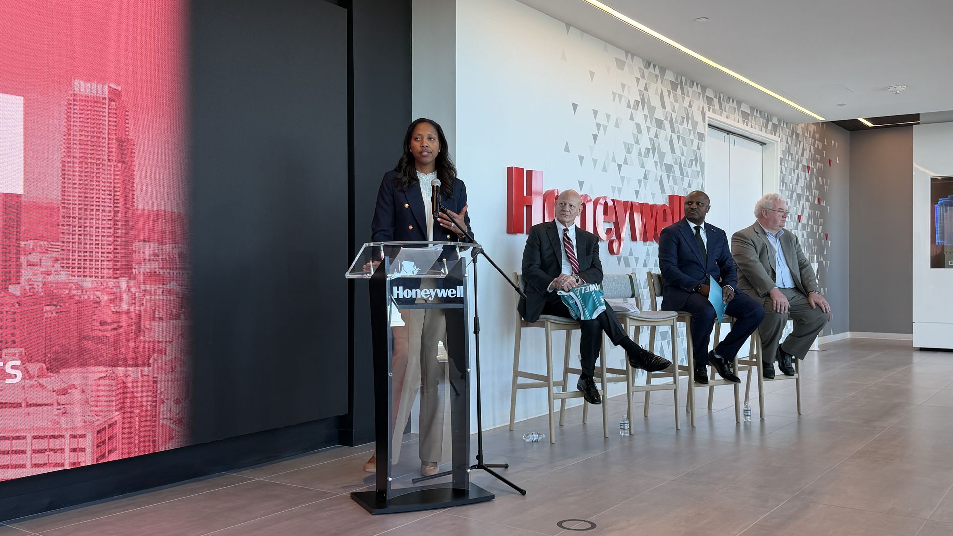 Hornets Sports & Entertainment president of business operations Shelly Cayette-Weston speaks at a Honeywell podium with microphone. Three men in suits sit on high chairs next to her against a white wall with red Honeywell logo.