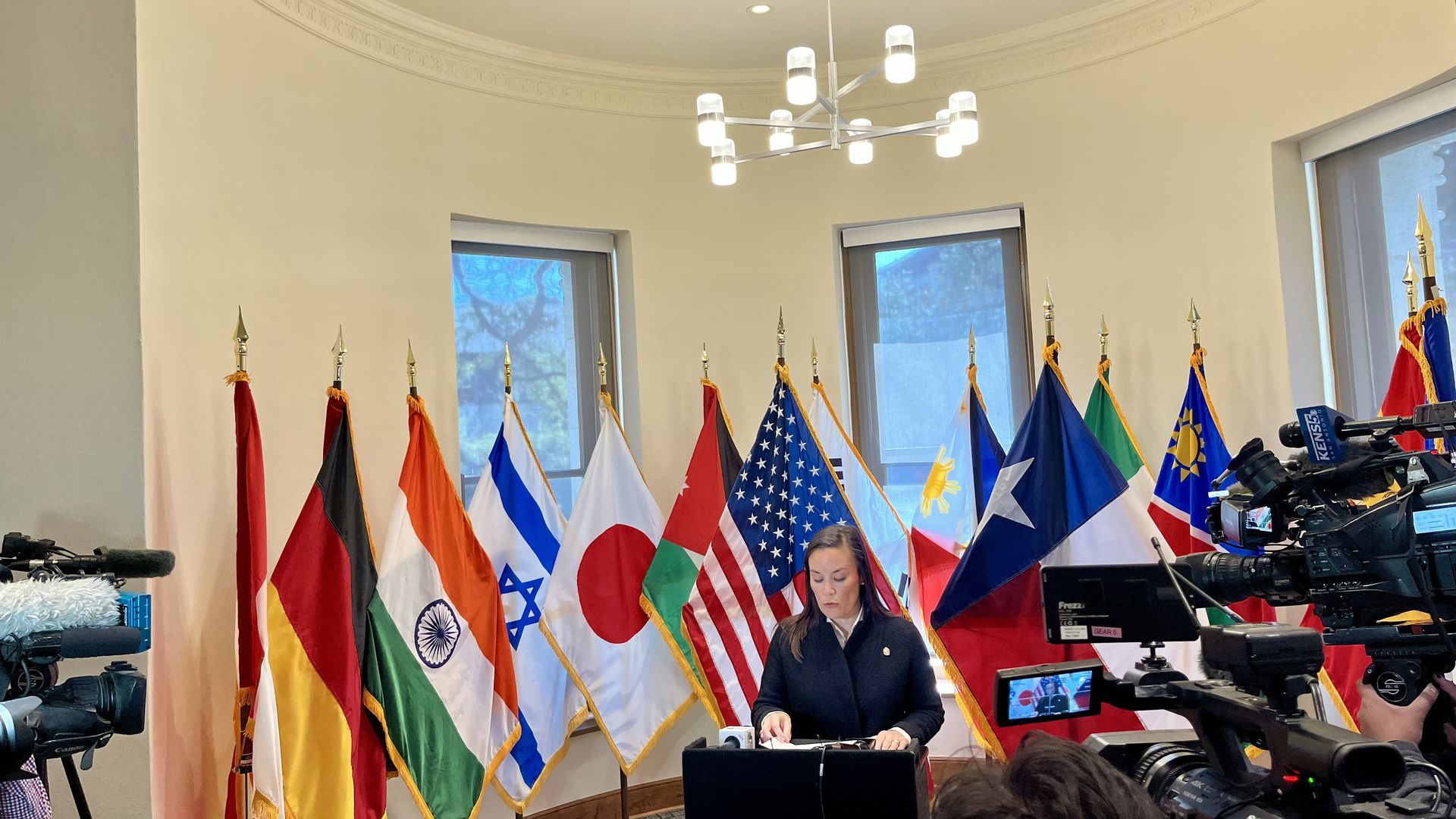 San Antonio Mayor Gina Ortiz Jones stands at a podium in a room with 11 international flags behind her. Cameras and microphones face her in a press setting.