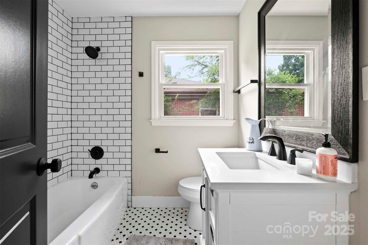 Bright bathroom with white subway-tile shower, black fixtures, hexagonal black-and-white floor tiles, white vanity, large black-framed mirror, and a window showing greenery outside.