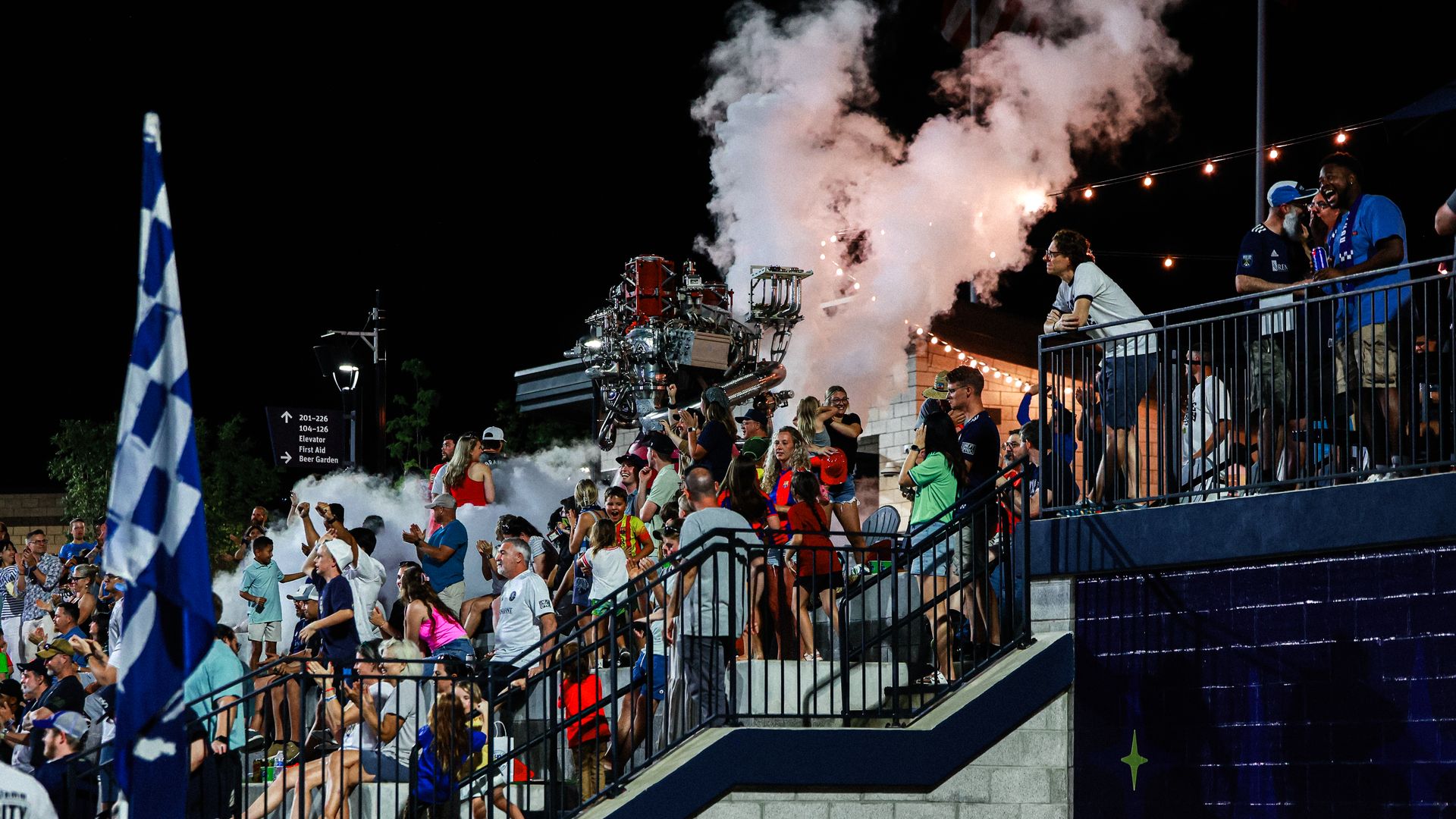 Crowd at a nighttime Huntsville City Football Club soccer match event cheering near a replica rocket engine emitting white smoke and sparks, with string lights overhead and a blue and white checkered flag in the foreground.