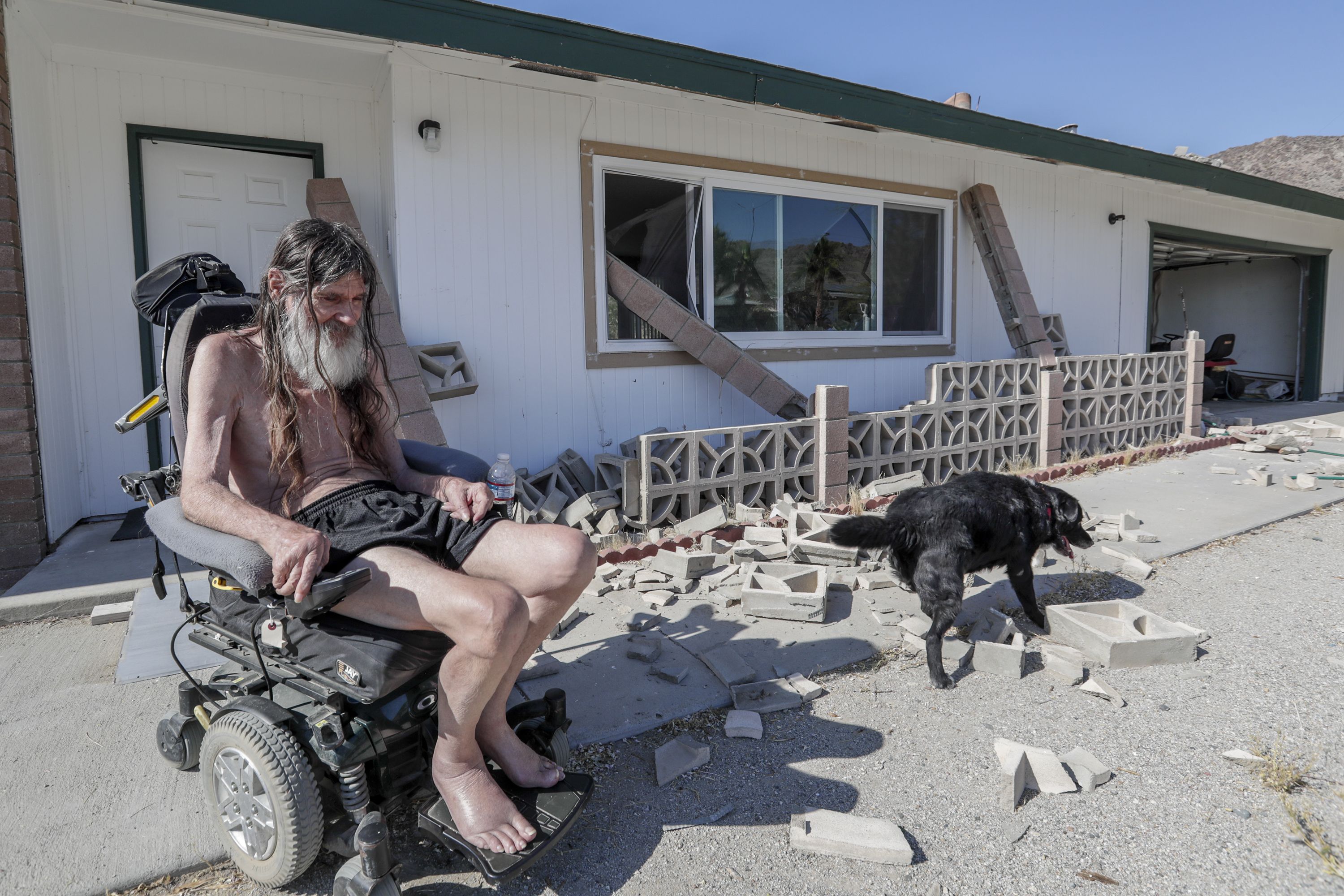 This image shows a shirtless man in an electric wheelchair and his dog in debris