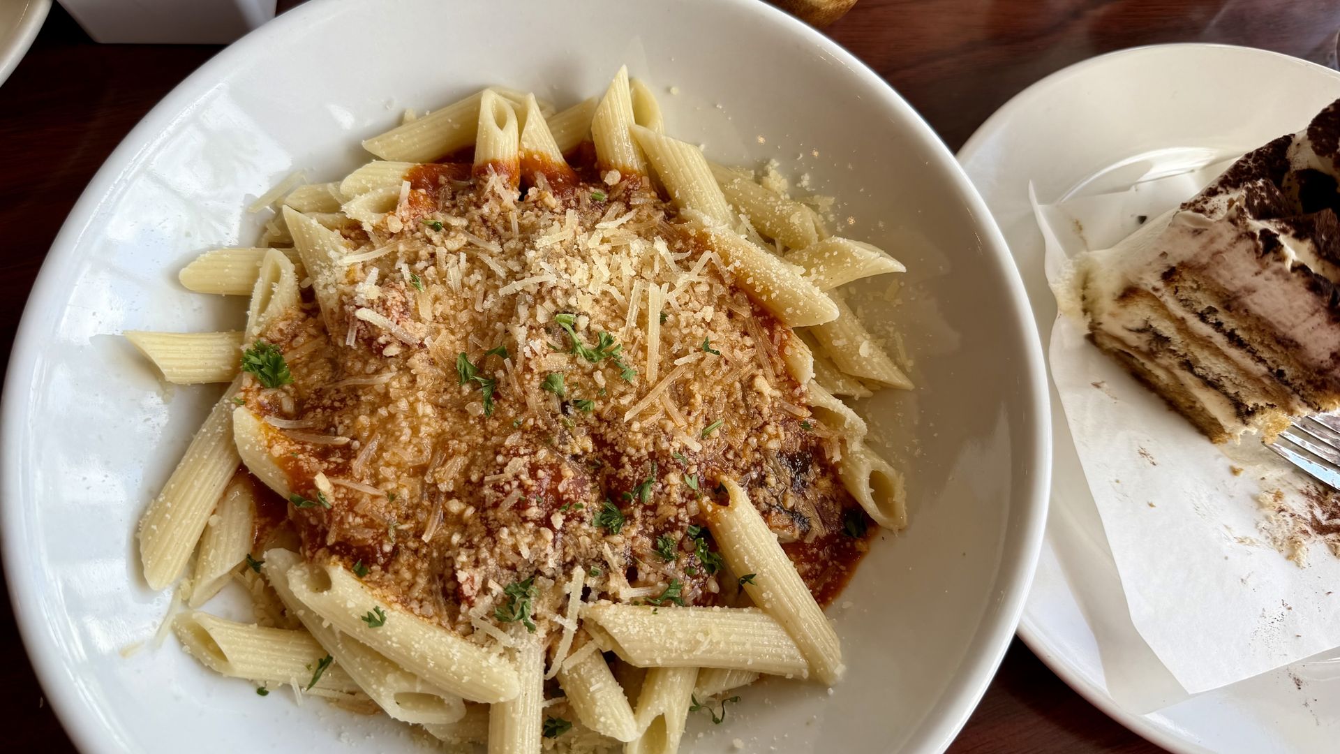 Top-down view of a white bowl of penne with tomato sauce, grated cheese, and parsley; a tiramisu slice on a plate sits beside a bread basket on a wooden table.