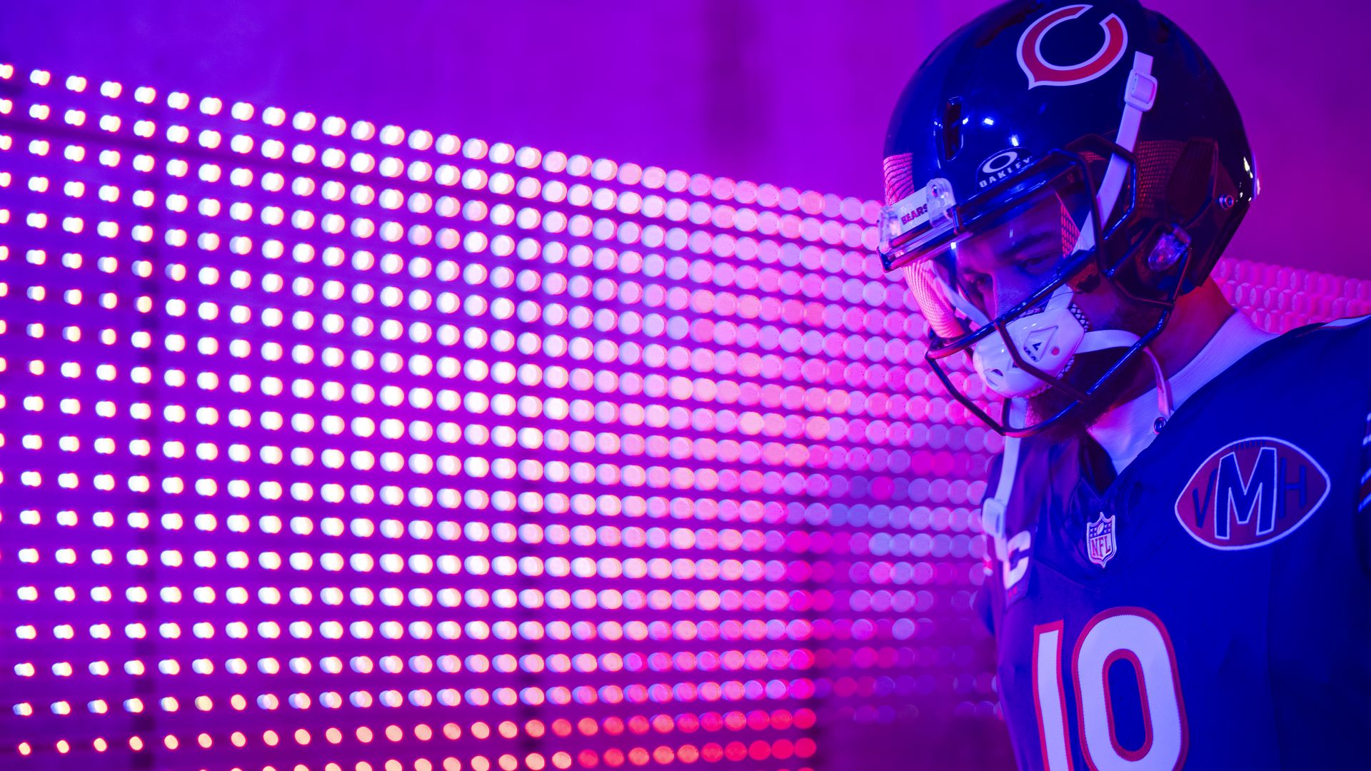 Close-up of a football player in a blue Chicago Bears jersey, wearing a dark helmet with a red-and-white 'C' logo, visor, and white chin strap, against a purple-pink LED light backdrop.