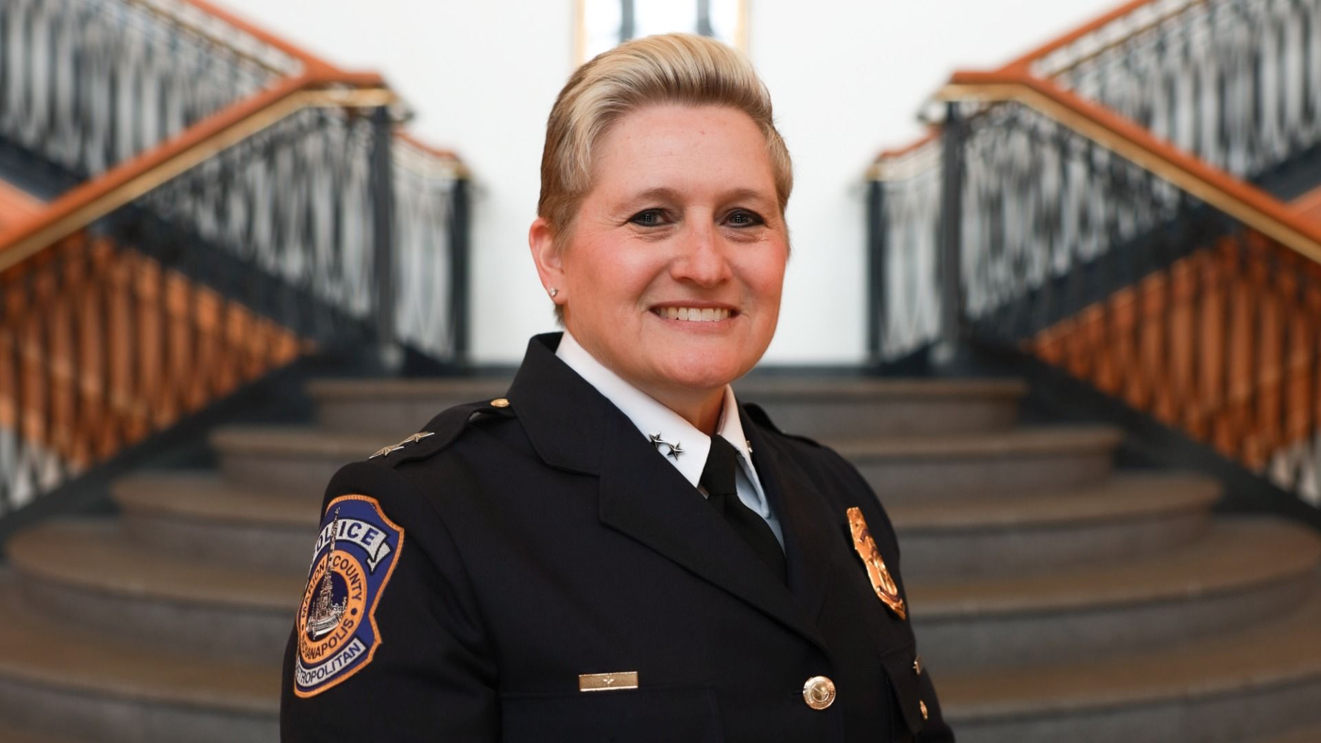 Smiling police officer with short blonde hair in black uniform with badges, standing in front of a staircase with black and wooden railings in a building.