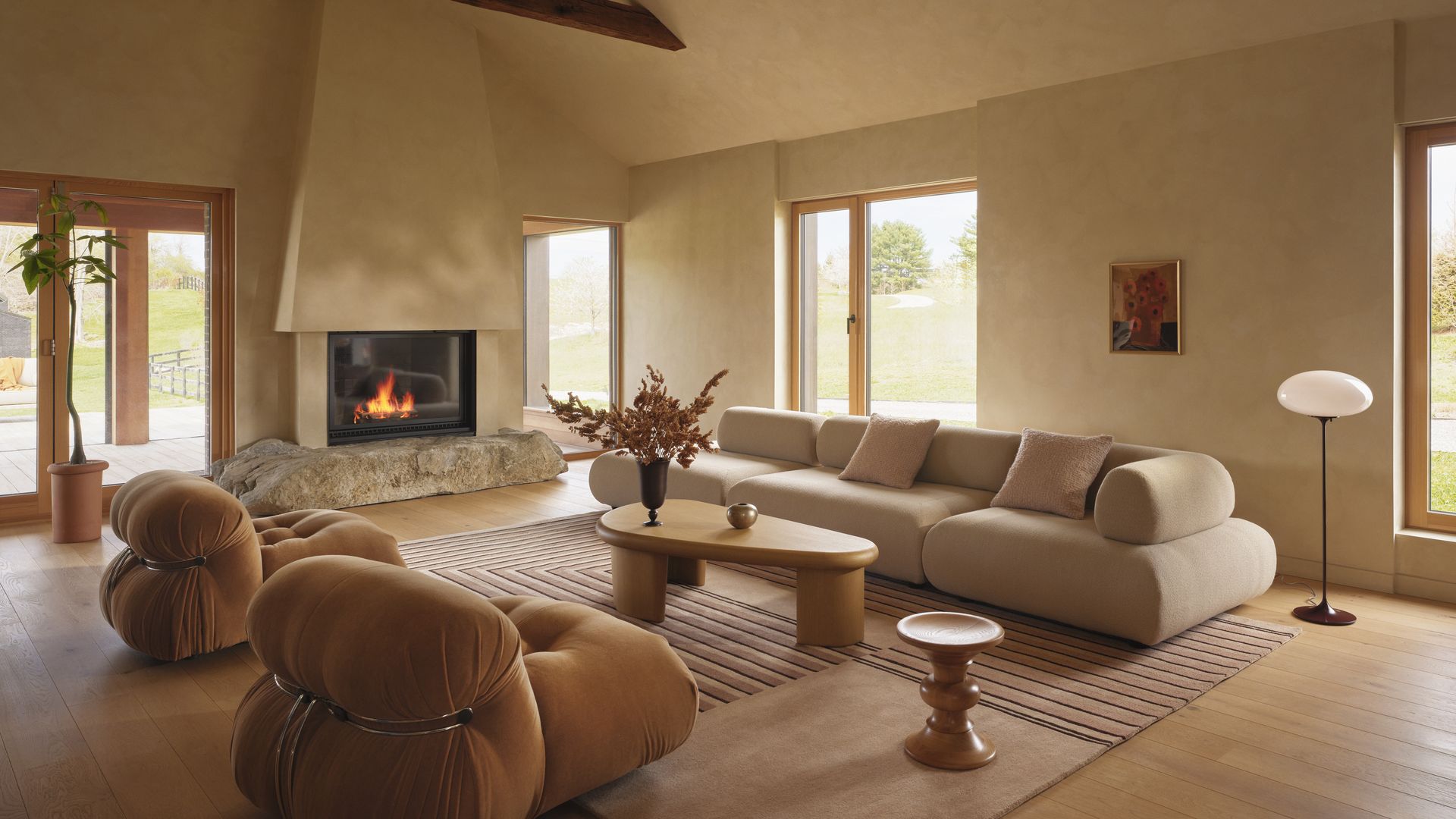 Warm beige living room with dark wooden ceiling beams, a stone-base fireplace, large windows, and a curved cream sofa with matching armchairs and a round wooden coffee table.