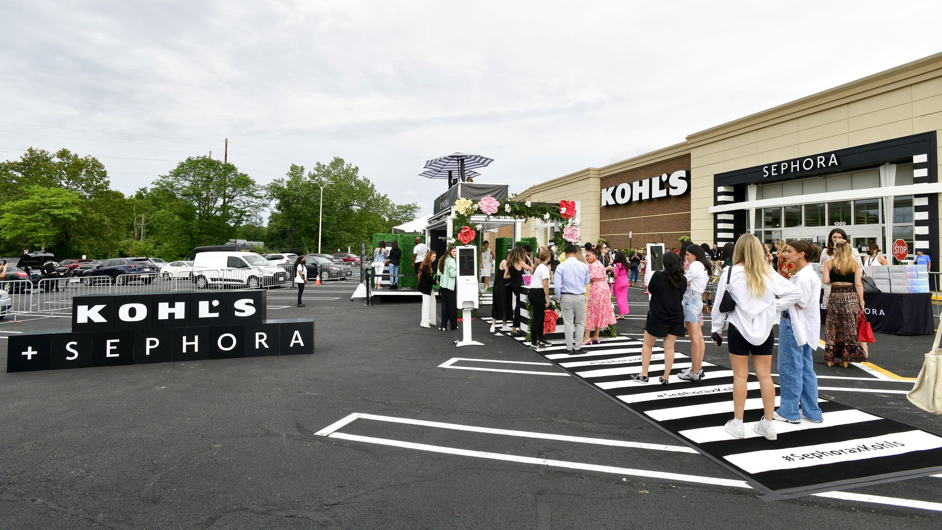 A crowd gathers in a freshly-paved black-asphalt parking lot in front of a Kohl's department store for a Sephora beauty store launch.