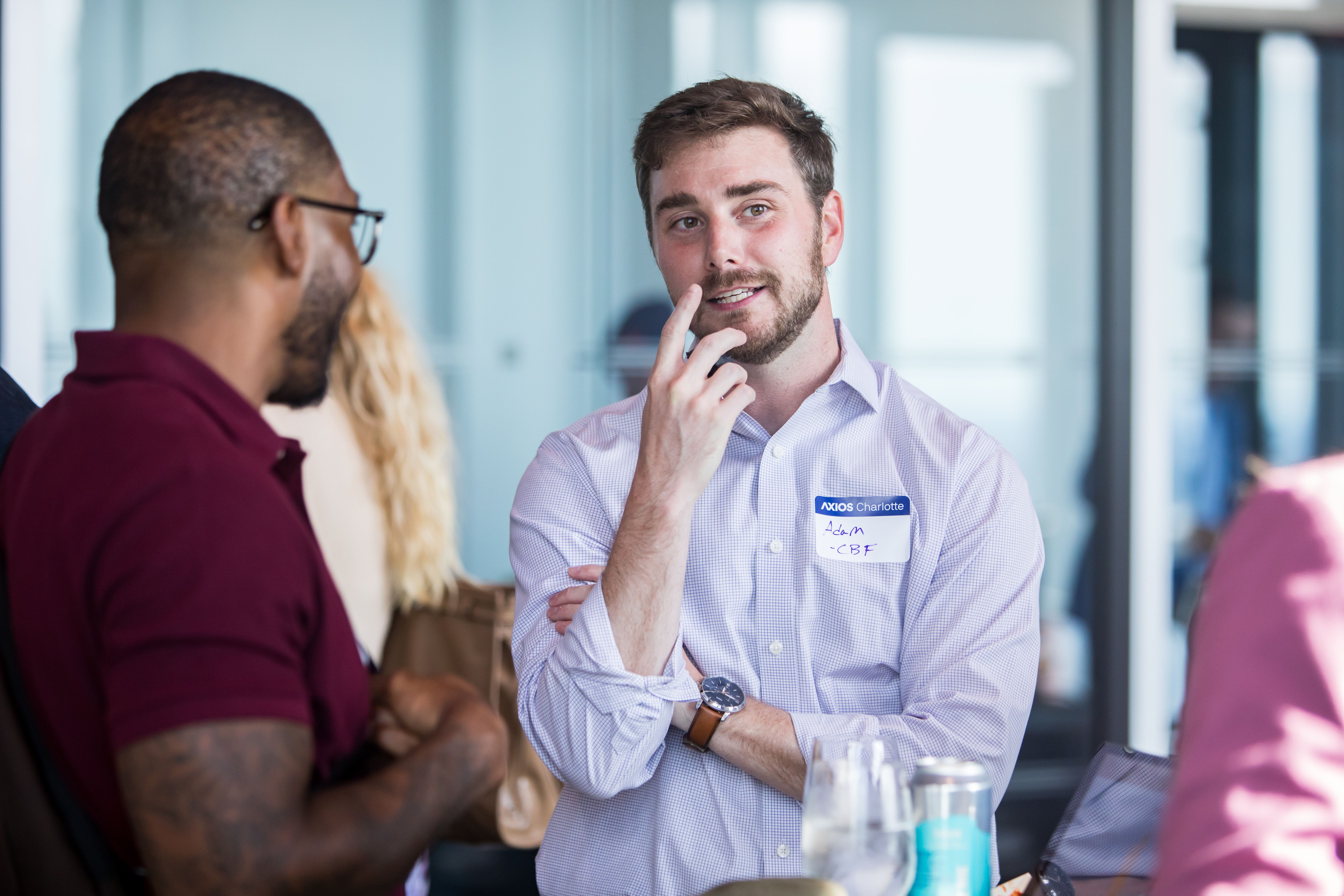 Man in a light purple checkered shirt with a name badge chats with others in a bright indoor setting, touching his chin as he speaks; others are blurred in the background.