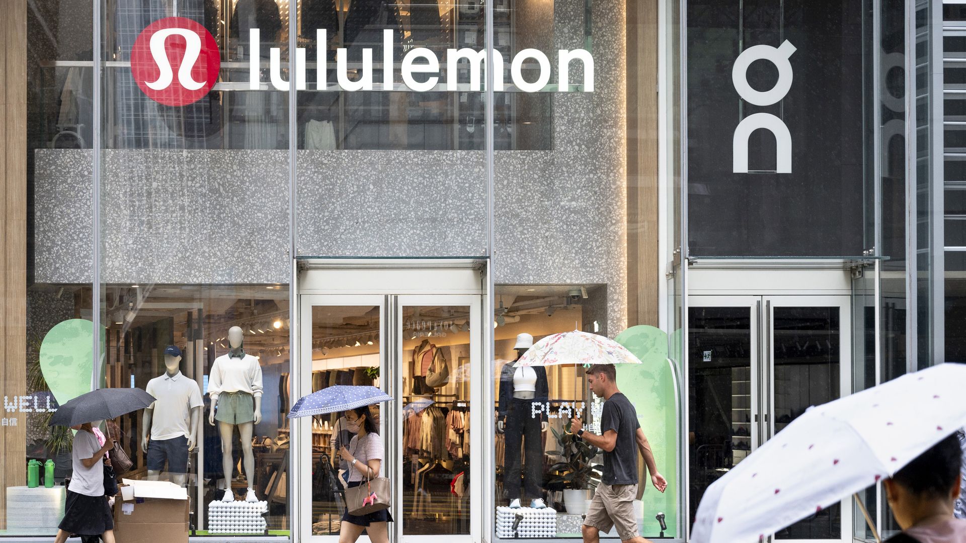 Pedestrians holding umbrellas walk by a Lululemon store in an urban setting.