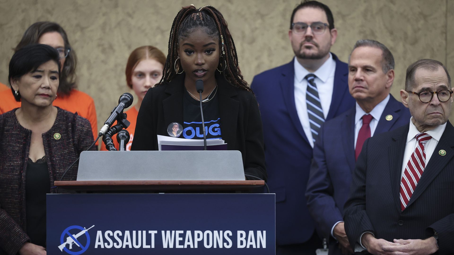 A Black girl with long hair stands at a podium that has a sign saying "Assault weapons Ban."