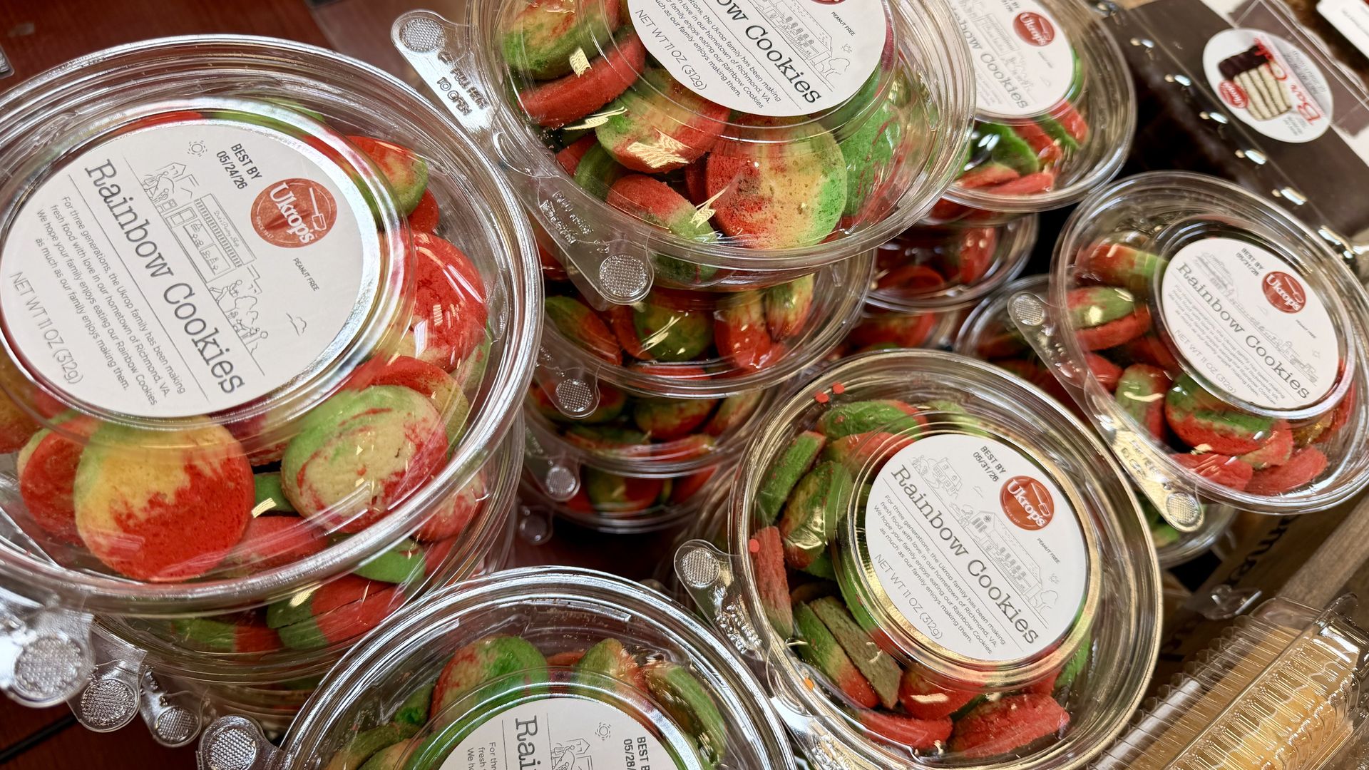 Clear plastic containers filled with red, green, and cream rainbow cookies; white labels read "Rainbow Cookies" with a red "Ukrop's" logo, stacked on a display.