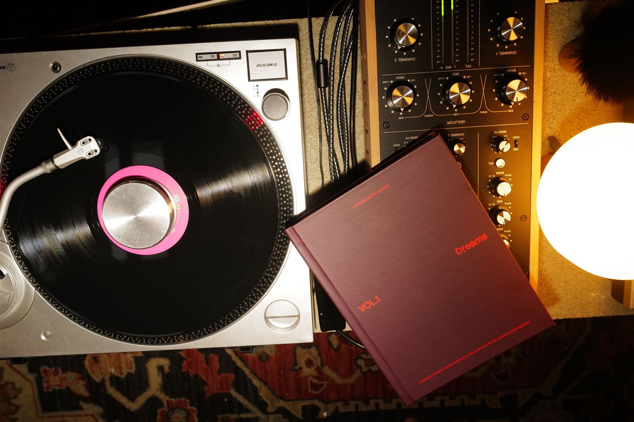 Top-down view of a DJ setup: a vinyl turntable with a pink slipmat and tonearm, a black mixer with gold knobs, tangled cables, a maroon notebook titled Dreams, and a white lamp on a patterned rug.